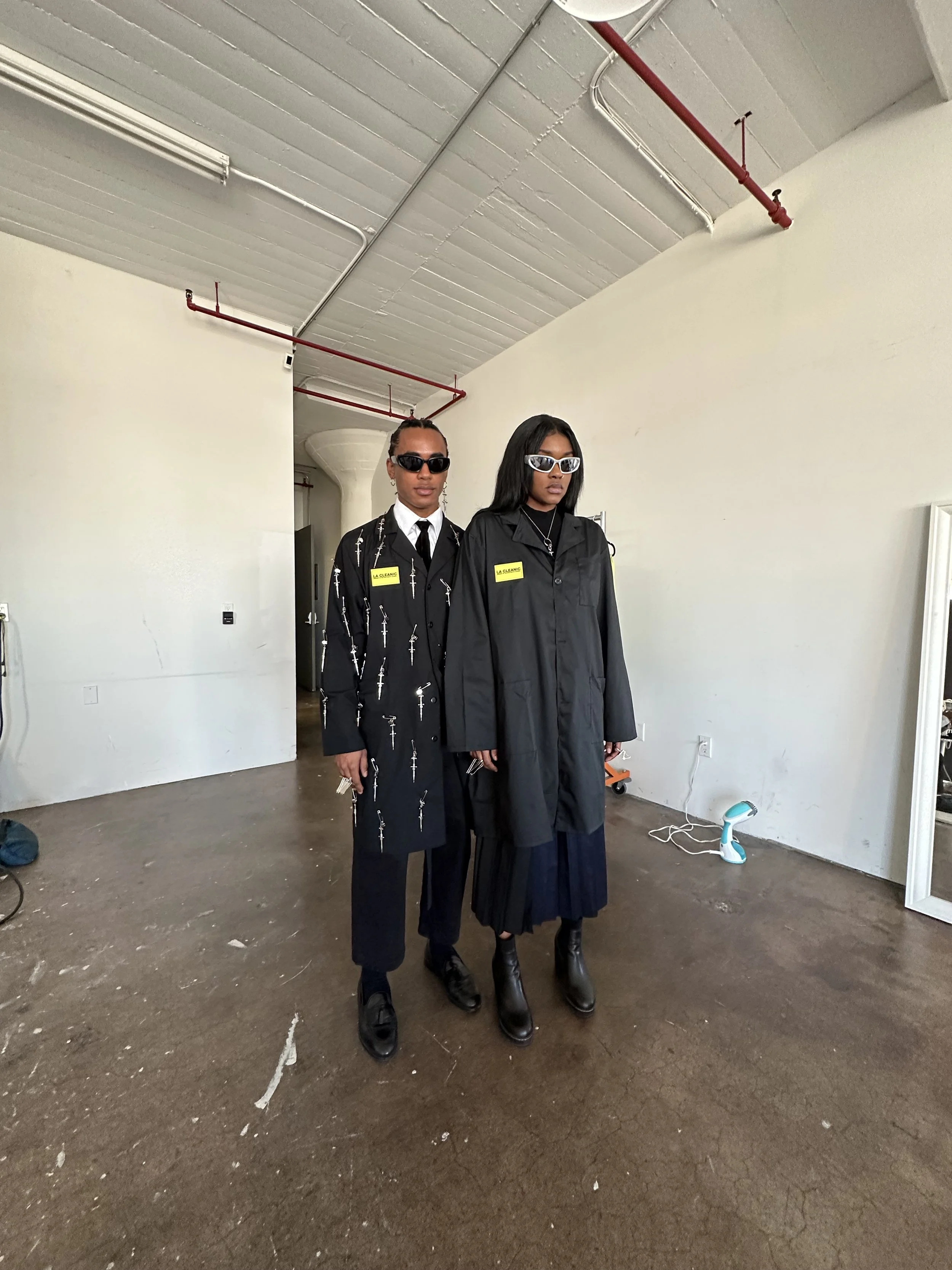 Two women standing side by side indoors, both wearing black outfits and sunglasses, with yellow name tags on their chests.