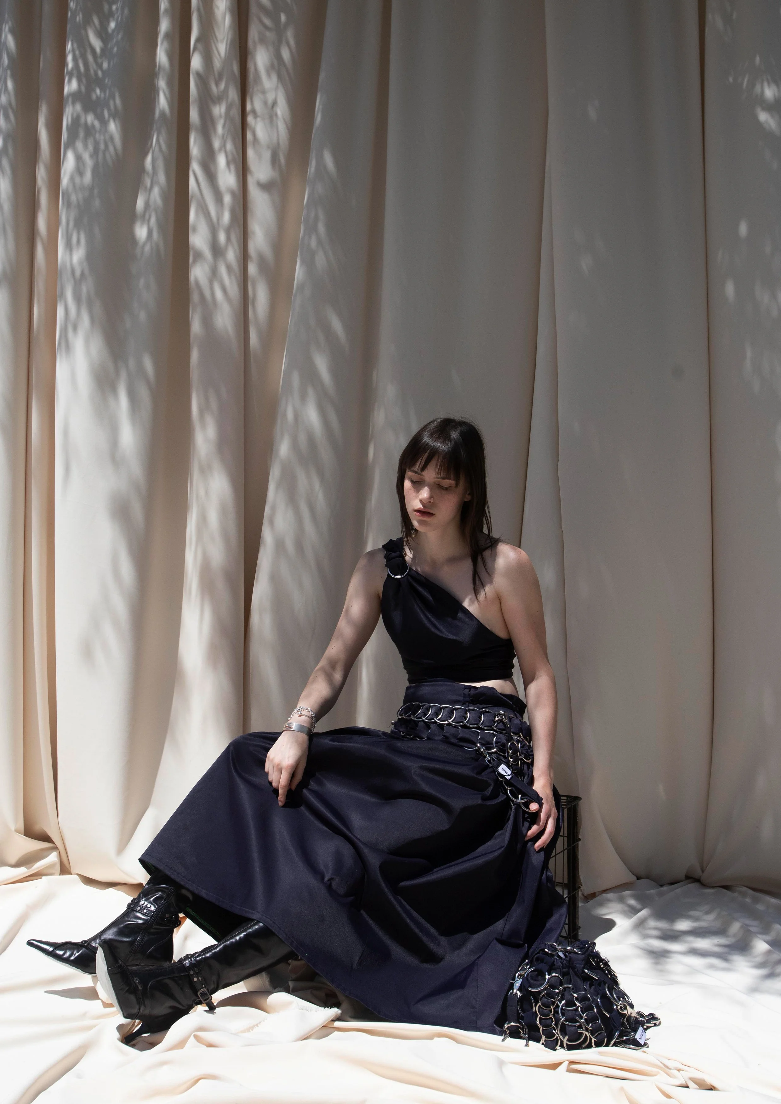 A young woman with dark hair and pale skin sits on the floor in front of light-colored curtains with shadow patterns. She wears a black asymmetrical top, a long black skirt with silver chain accessories, and black knee-high boots. She has silver jewelry and a black handbag on the floor beside her.
