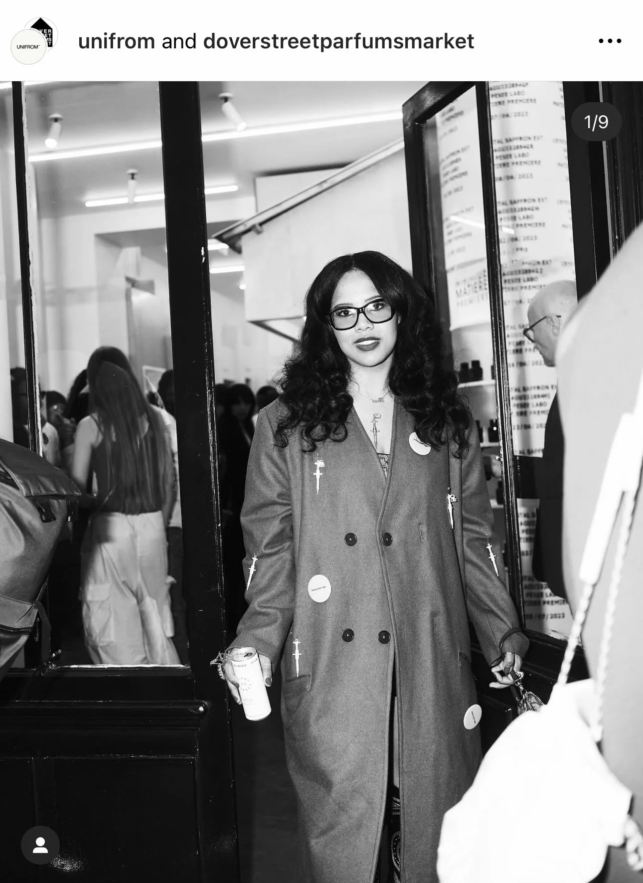 A woman with long, curly hair, glasses, and dark lipstick standing at a store entrance, holding a beverage can, surrounded by people in the background.