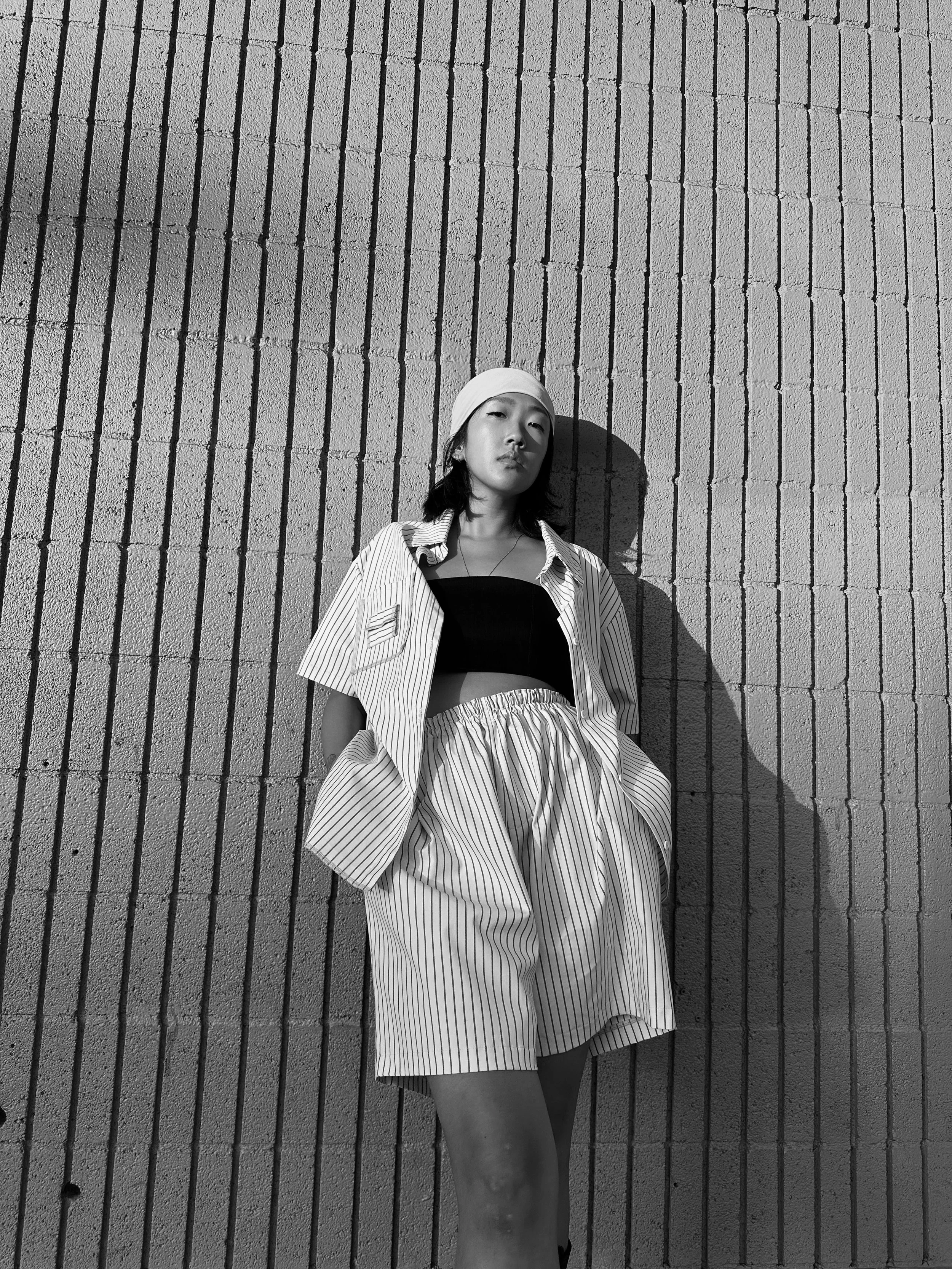 A young woman with shoulder-length dark hair wearing a headscarf, a striped shirt over a black crop top, and striped shorts, standing against a textured brick wall with her hands in her pockets.