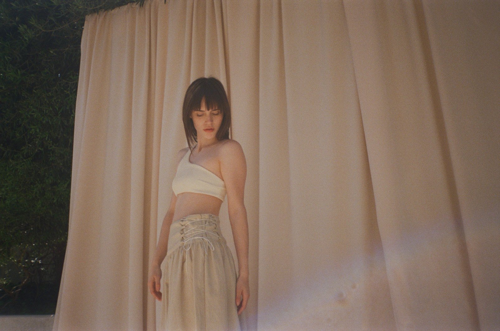 A young woman with dark hair and bangs wearing a white crop top and a long beige skirt, standing beside beige curtains, looking downward with a neutral expression.