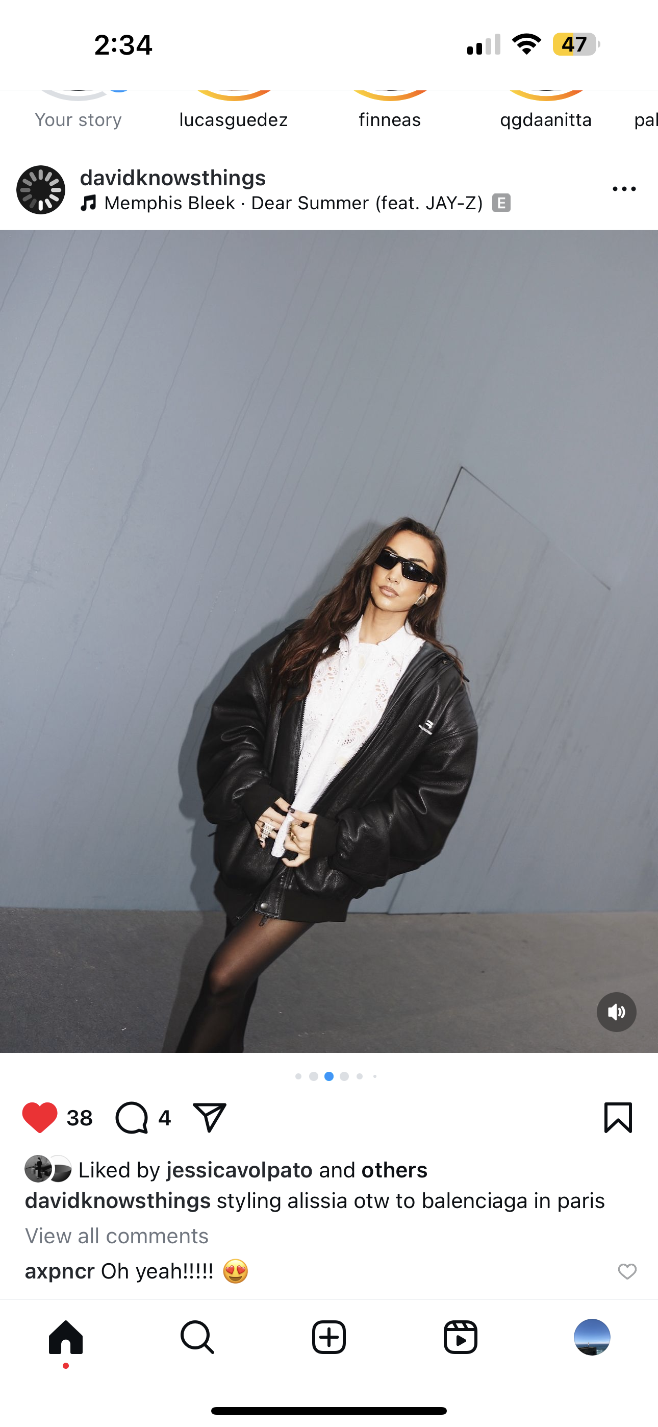 A woman standing against a gray wall, wearing black sunglasses, a white top, a black leather jacket, and black tights.