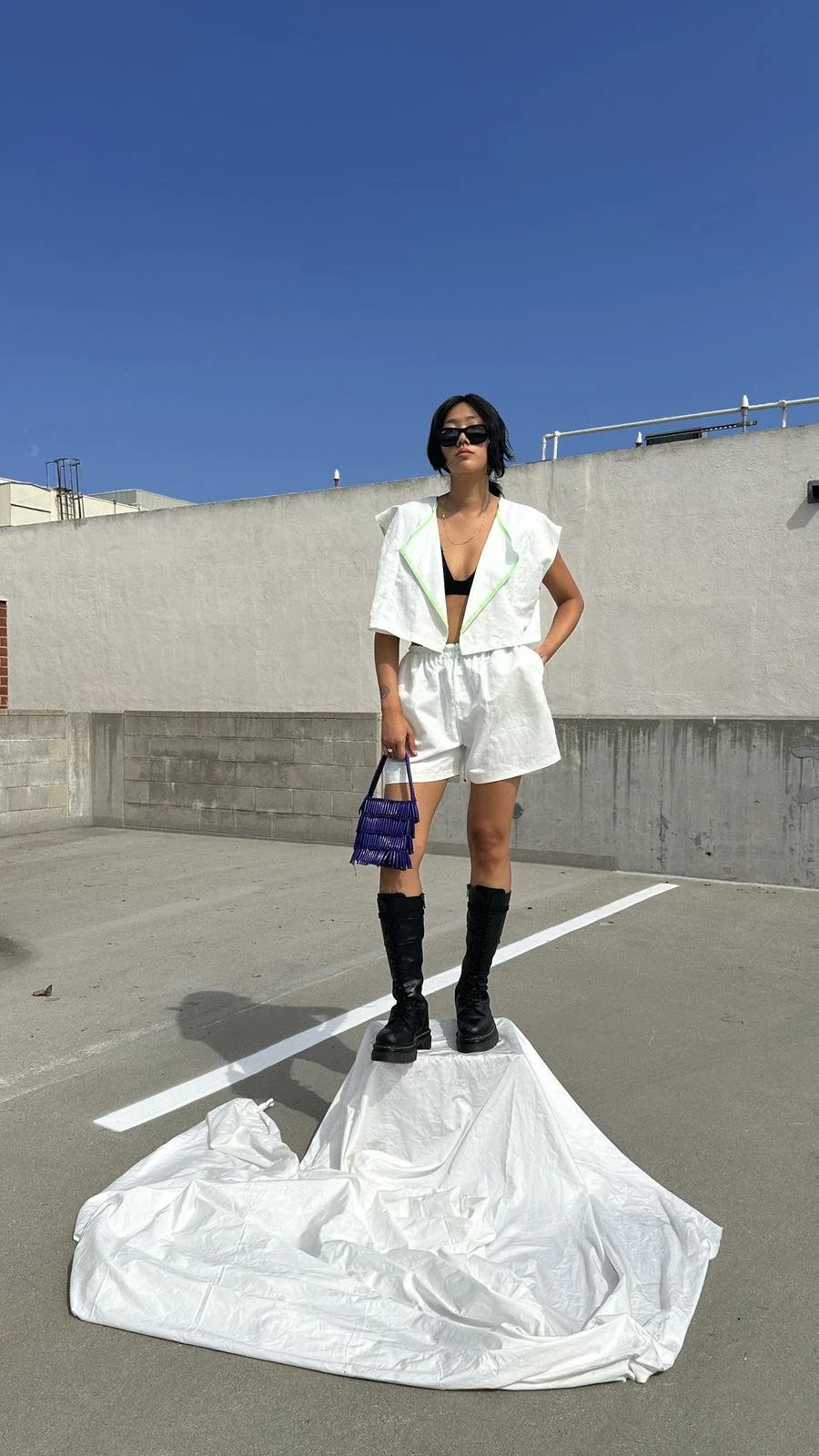 A woman standing on a white cloth on a parking lot, wearing black sunglasses, a black sports bra, white shorts, a white vest, and black boots, holding a purple handbag, against a blue sky and plain wall.