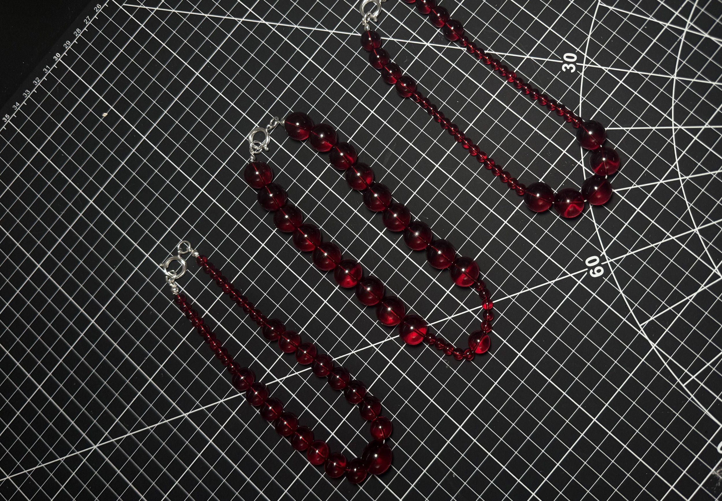 Red beaded jewelry, including a necklace and a bracelet, laid on a black cutting mat with white grid lines and measurement markings.