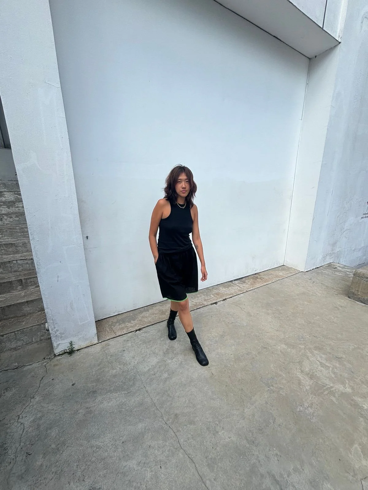 Young woman with wavy hair wearing a black tank top, black shorts, and black boots walks in front of a white wall.
