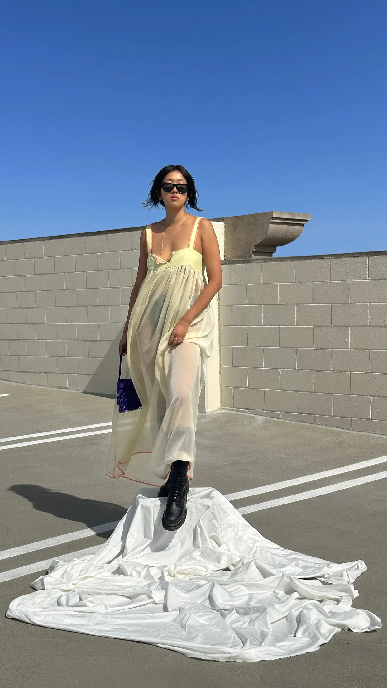 A woman standing on a crumpled white fabric on a parking lot, wearing a light yellow dress, black combat boots, and sunglasses, with a blue sky in the background.