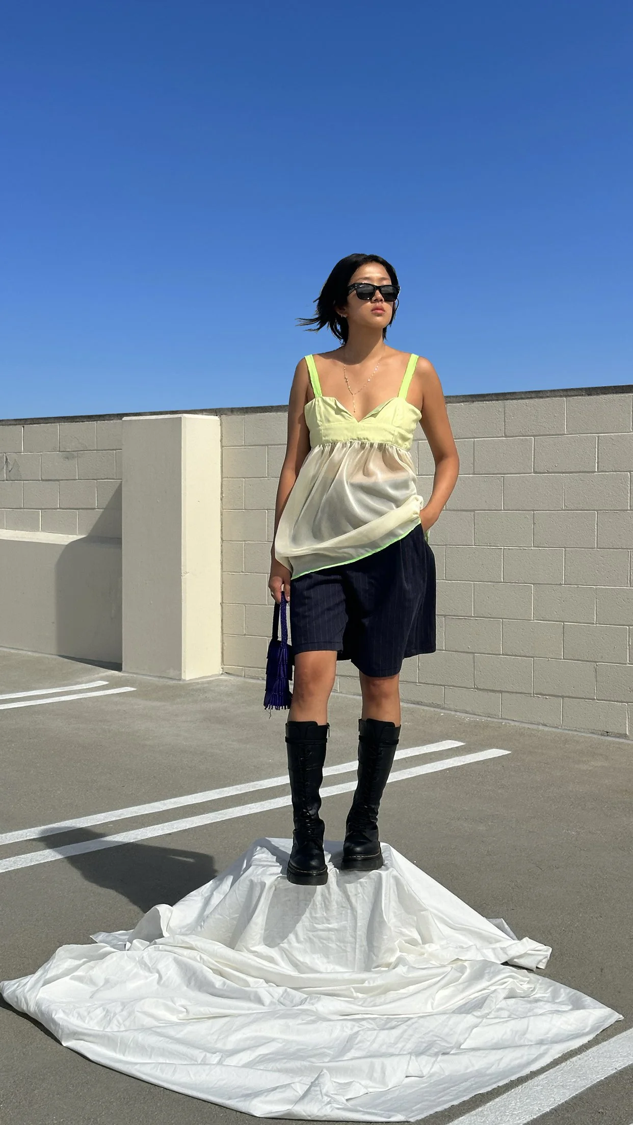 A woman with black hair, sunglasses, and a yellow top standing on a white fabric sheet on a parking lot, with a beige brick wall and a clear blue sky in the background.
