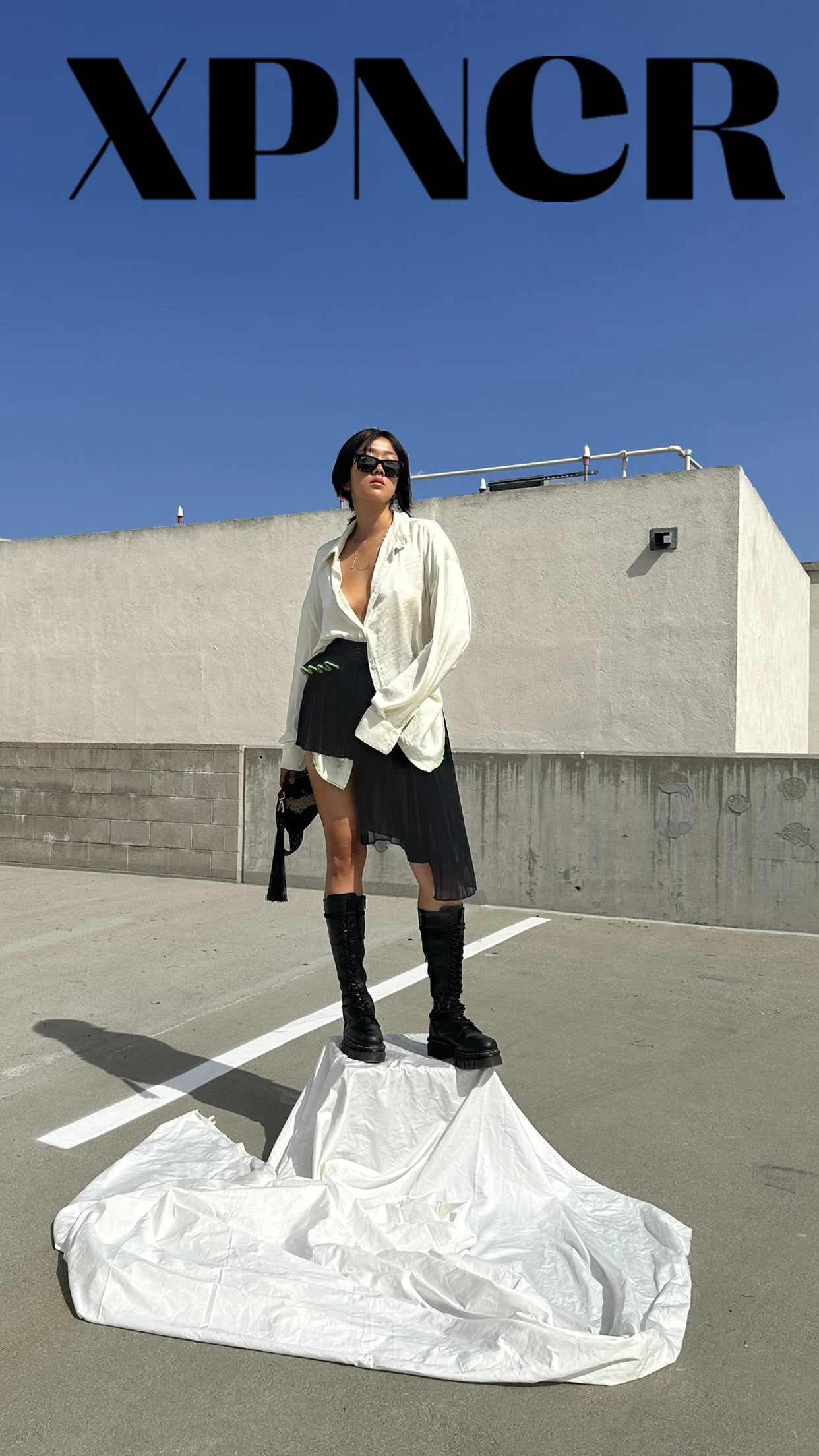 A woman standing on a makeshift white fabric sculpture in an outdoor parking lot with a blue sky background, wearing black boots, black skirt, white shirt, and sunglasses. The top of the image displays the text 'XPNCR' in large black letters.