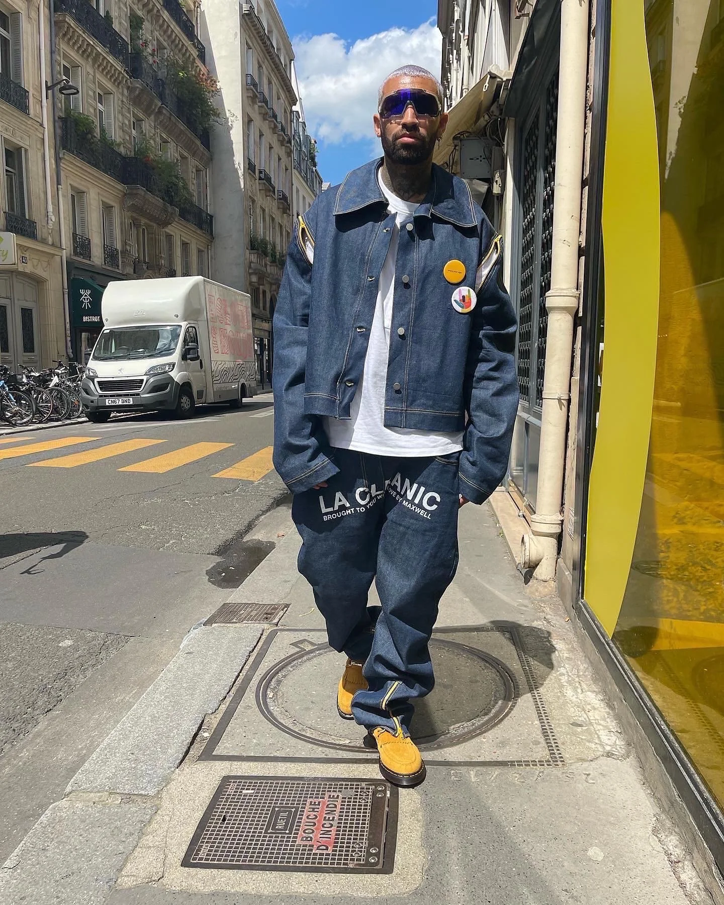 A man wearing sunglasses, a denim jacket with pins, a white t-shirt, baggy denim jeans, and yellow shoes walking on a city sidewalk with buildings, cars, and bicycles in the background.