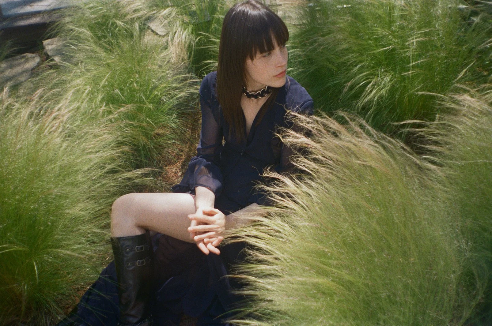 A young woman with dark hair and bangs sitting amidst tall green grass, wearing a navy blue dress, knee-high black boots, and a black choker necklace, looking to the side.