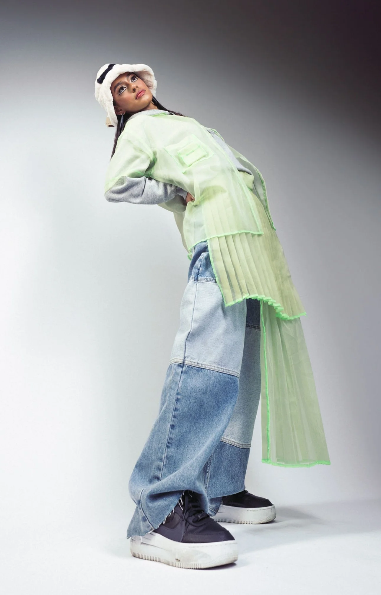 A young woman posing in an edgy fashion outfit, wearing a panda hat, a neon green transparent jacket, oversized jeans, and sneakers.