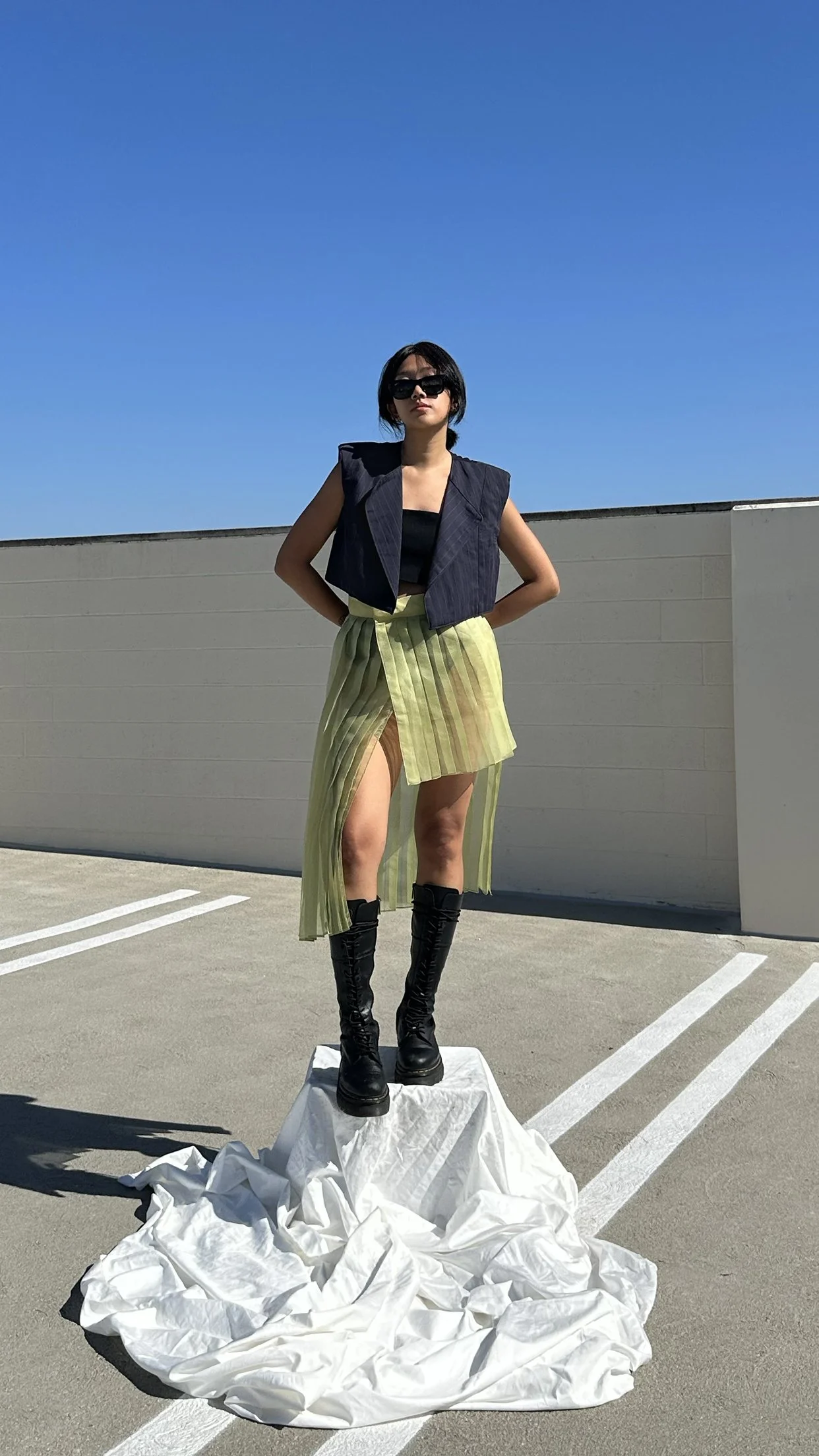 A woman standing on a small pedestal covered with white fabric in a parking lot against a blue sky, wearing black sunglasses, a black top, a sleeveless dark vest, a yellow-green pleated skirt with a high slit, and black lace-up boots.