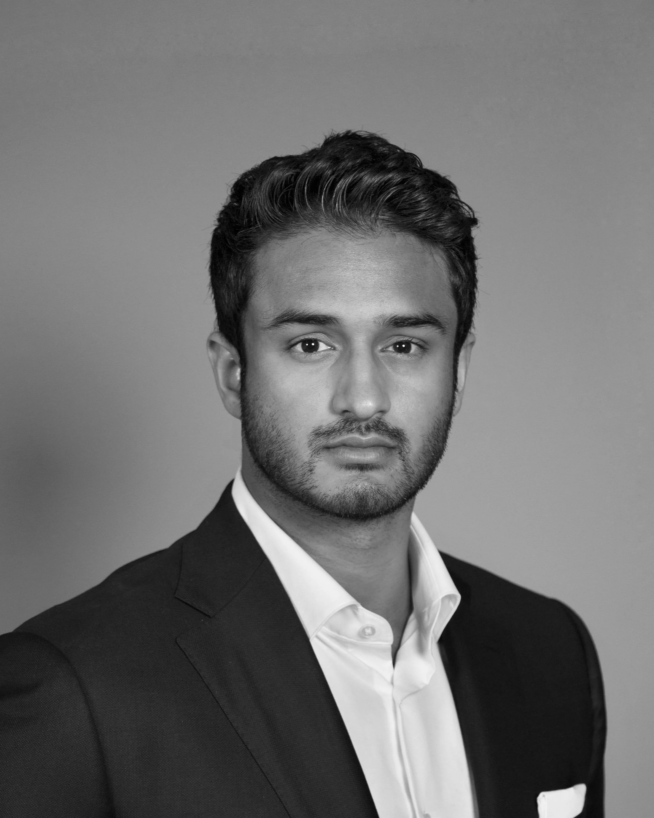 Black and white portrait of a young man with dark hair, wearing a suit jacket and shirt, against a plain background.
