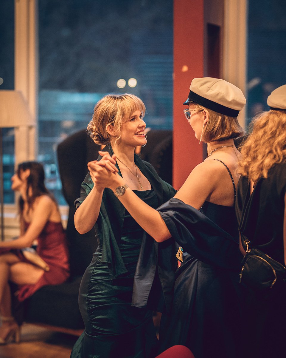 Två kvinnliga studenter dansar tillsammans på Chalmers Studentkårs höstbal bärandes studentmössor.  I bakgrunden dansar och minglar fler studenter.