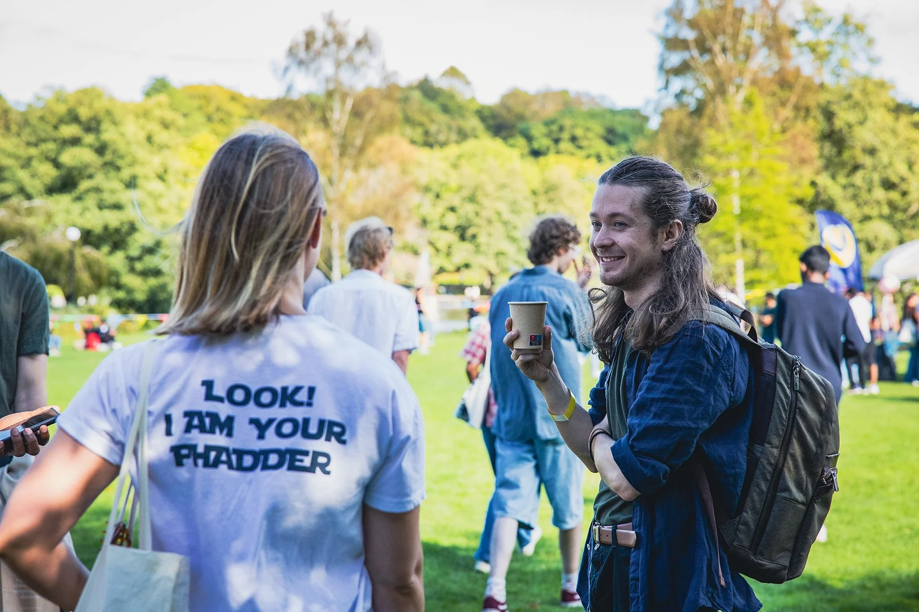 En man med långt hår, bärande ryggsäck, samtalar med en person i en vit t-shirt som har texten 'LOOK! I AM YOUR PHADDER' på ryggen, utomhus på en gräsmatta.