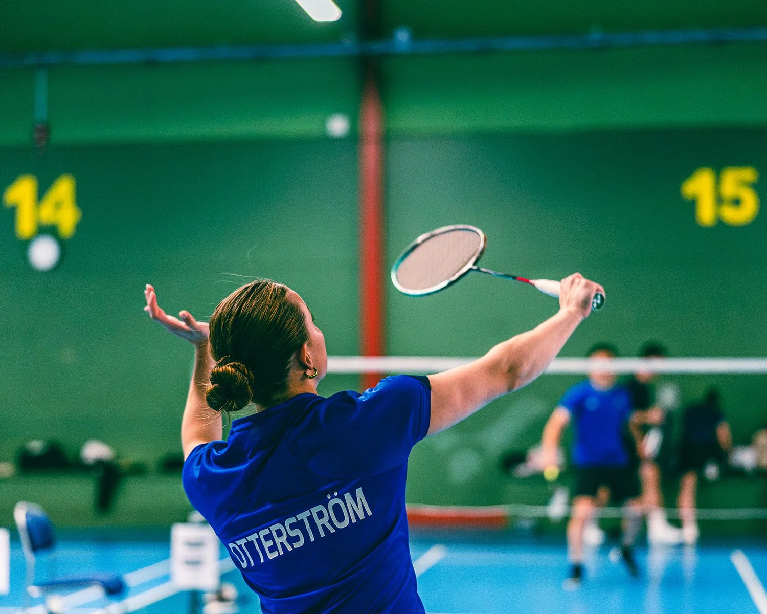 En kvinna spelar badminton inomhus, sedd bakifrån, på Akademiska Spelen.
