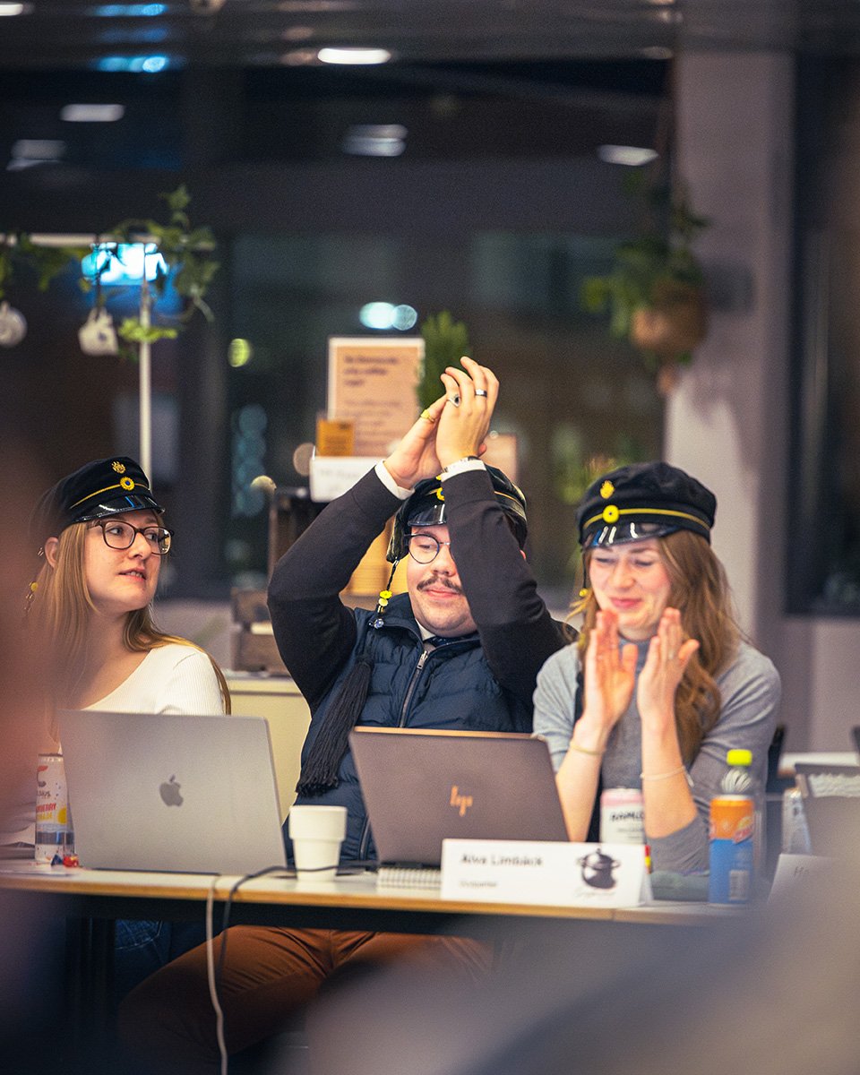 Tre personer i studentmössor firar, en man i mitten håller händerna högt ovanför huvudet medan två kvinnor på vardera sida ser glada ut, sittandes vid ett bord med laptops.