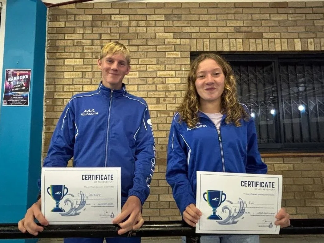 Two young individuals, a male and a female, smiling and holding certificates of achievement, stand in front of a brick wall in an indoor setting. They are both dressed in blue sports jackets with the AquAzurra logo.