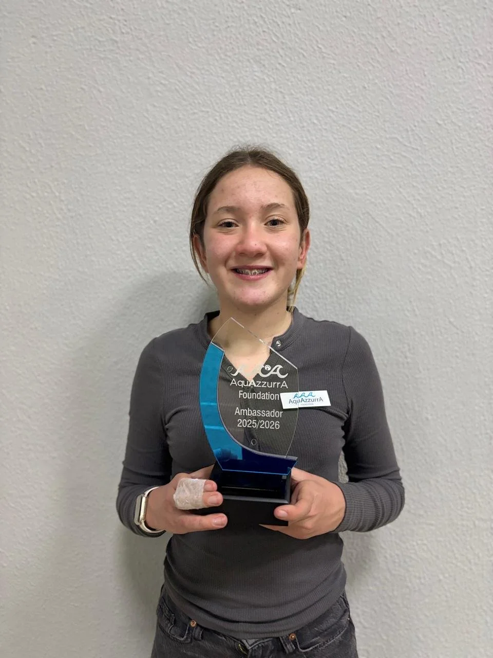 Young girl smiling and holding an award with a clear acrylic top, blue accent, and black base, standing against a plain off-white wall.