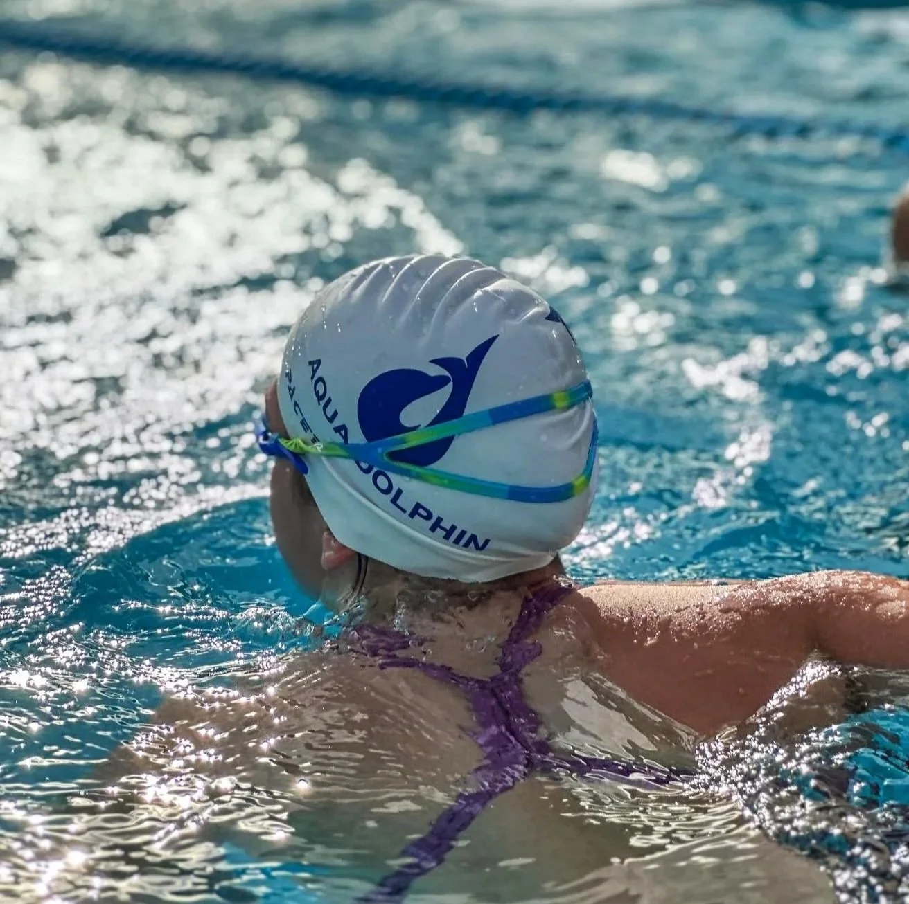 Swimmer wearing a white swim cap with the text 'Aqua Dolphin' and a dolphin logo, swimming in a pool with sunlight reflecting on the water.