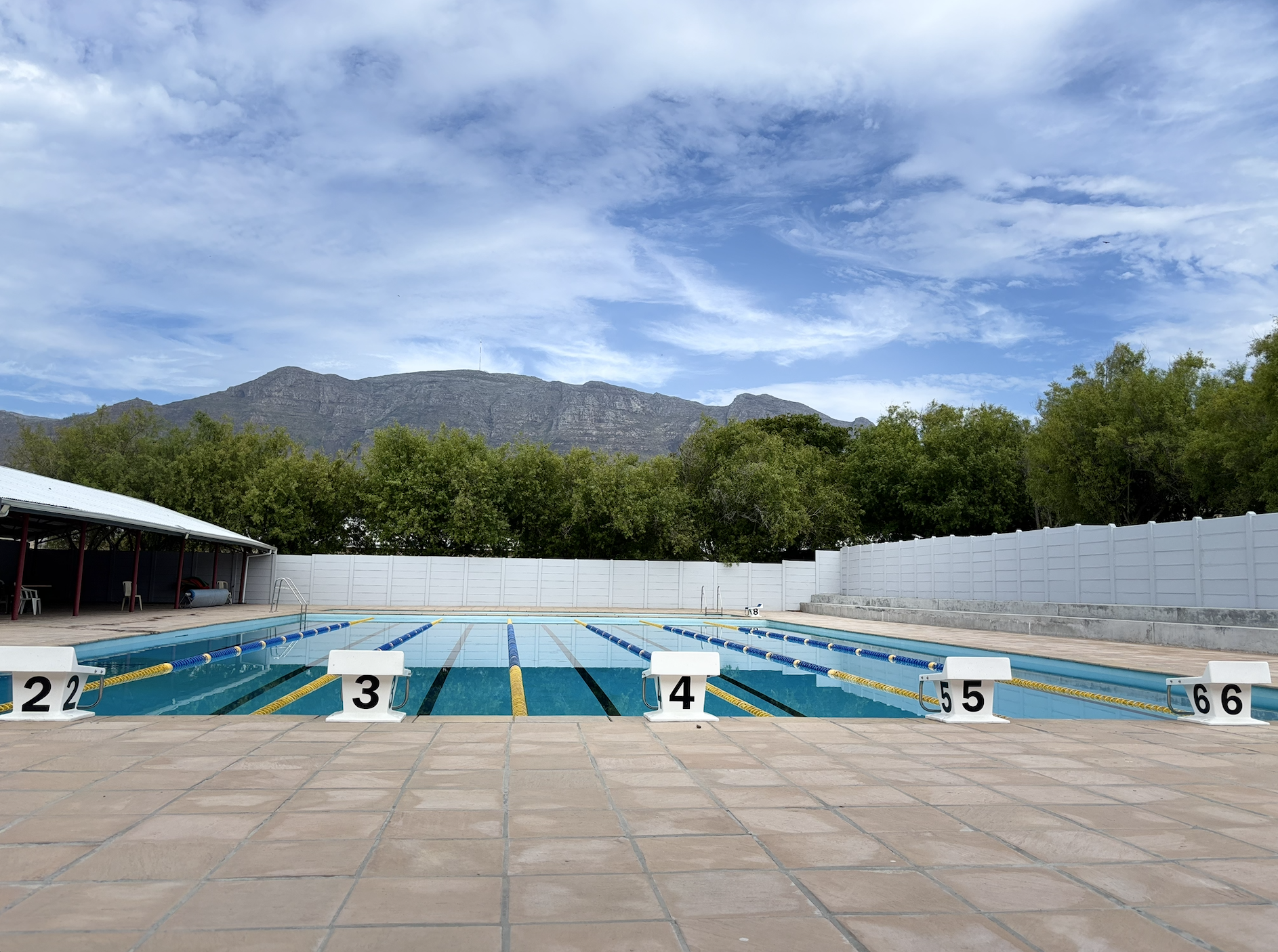 Empty outdoor swimming pool with lane dividers, poolside chairs under a shelter, white fence, lush green trees, mountain in the background, partly cloudy sky.