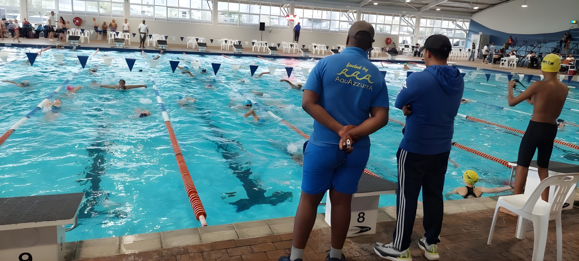Swimmers practicing or competing in an indoor swimming pool, with two officials or coaches standing on the pool deck watching, and others in the pool wearing swim caps.