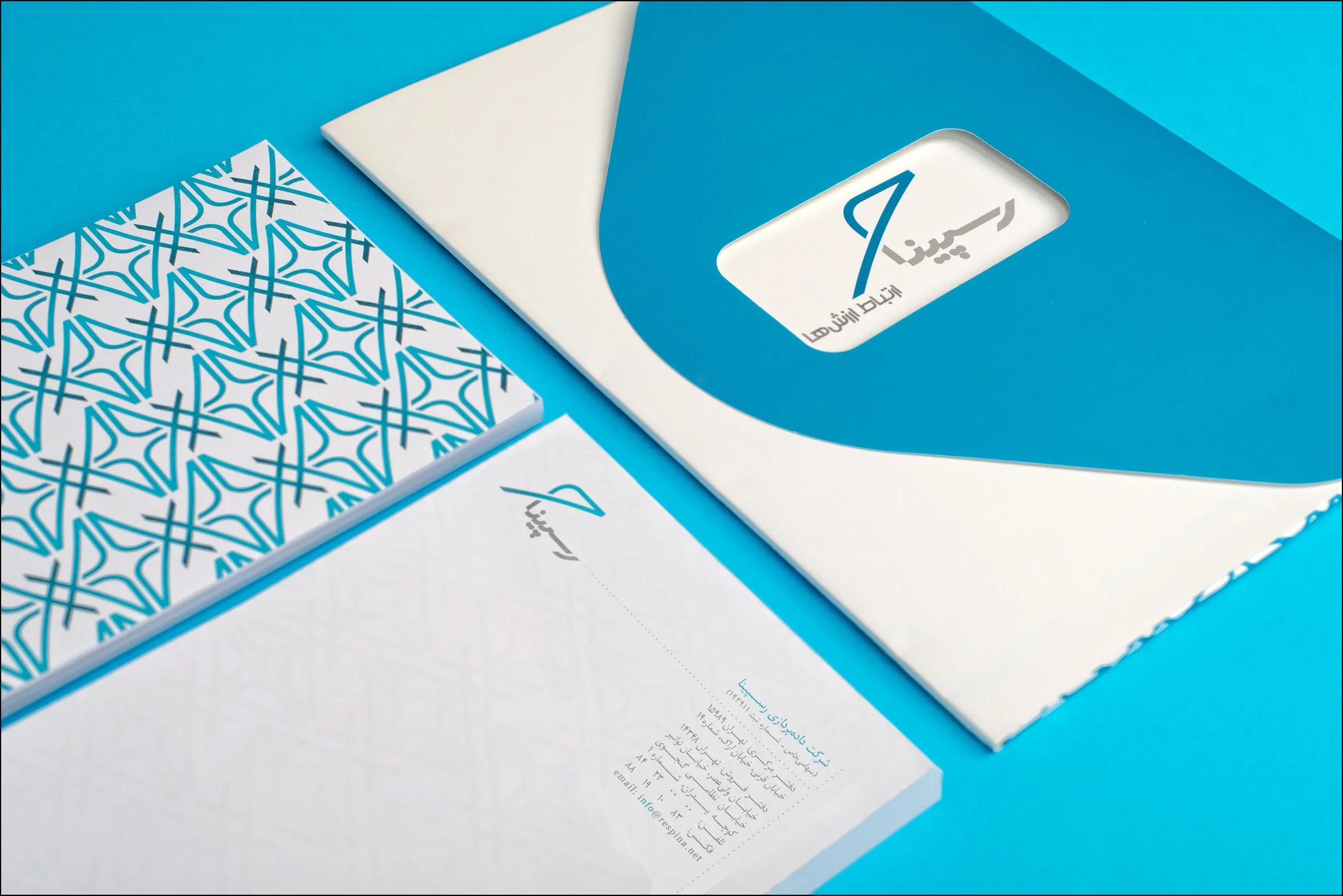 Collection of printed materials, including a blue folder with a rectangular cutout revealing a logo with Arabic text, a patterned notepad with blue and white geometric design, and a white sheet with Arabic text and contact information, all on a brigh