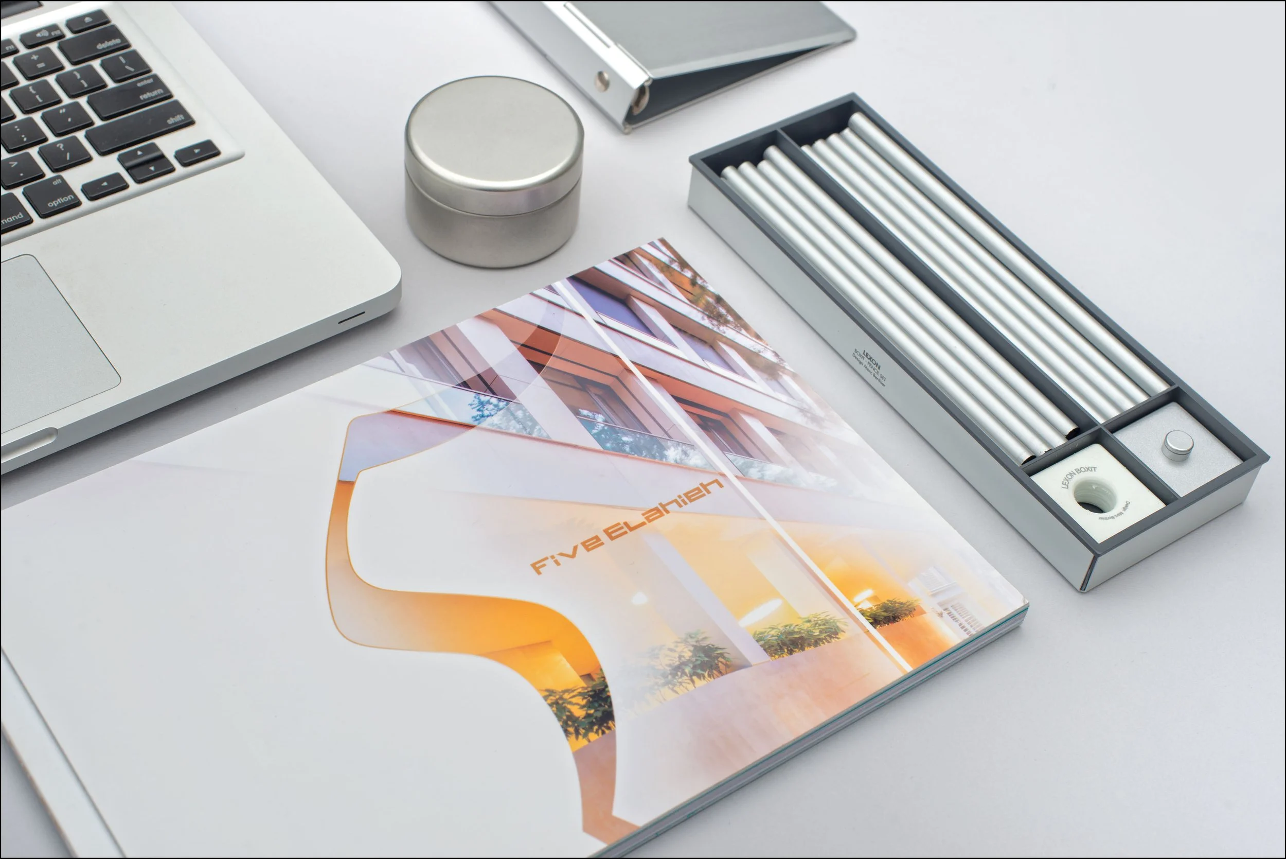 Open laptop, metallic container, notebook, box of white chalk sticks, and a brochure on a white desk.