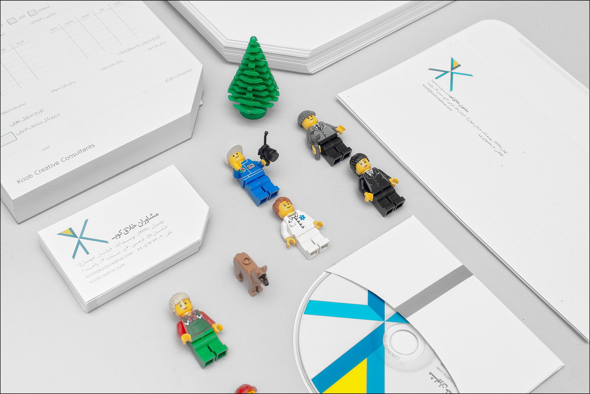 Arrangement of Lego figures, a green container tree, and printed materials, including business cards, stacks of paper, and a CD, on a white surface.