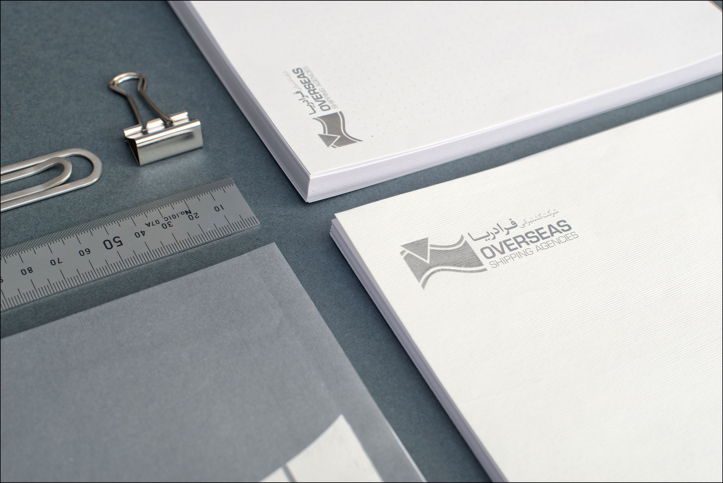 Business stationery featuring Oversaes Shipping Agencies logo, a notepad, ruler, binder clip, and paper clips on a grey surface.