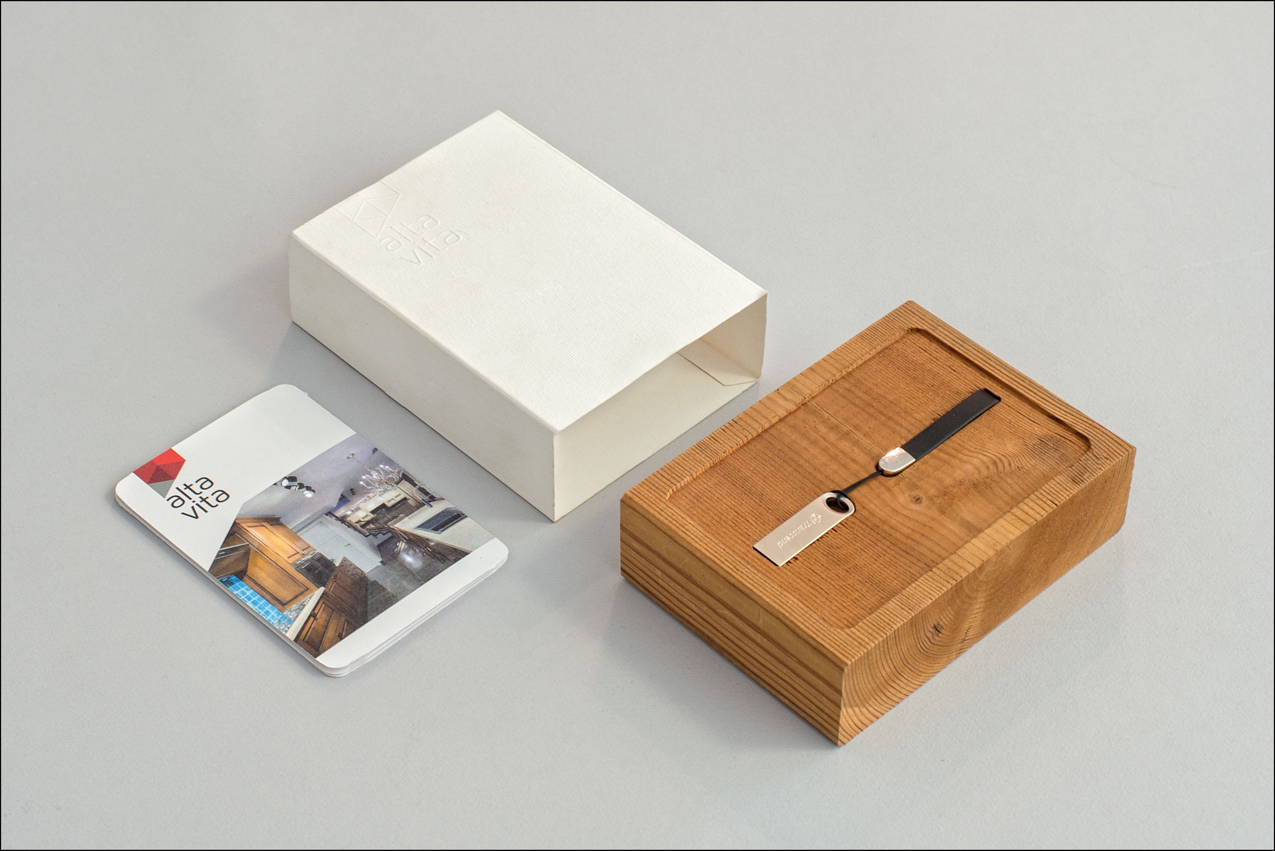 A set of Alta Vista branded items including a white cardholder, a brochure or flyer, a wooden box with a leather strap, and a metal keychain with a leather strap, all arranged on a light gray surface.