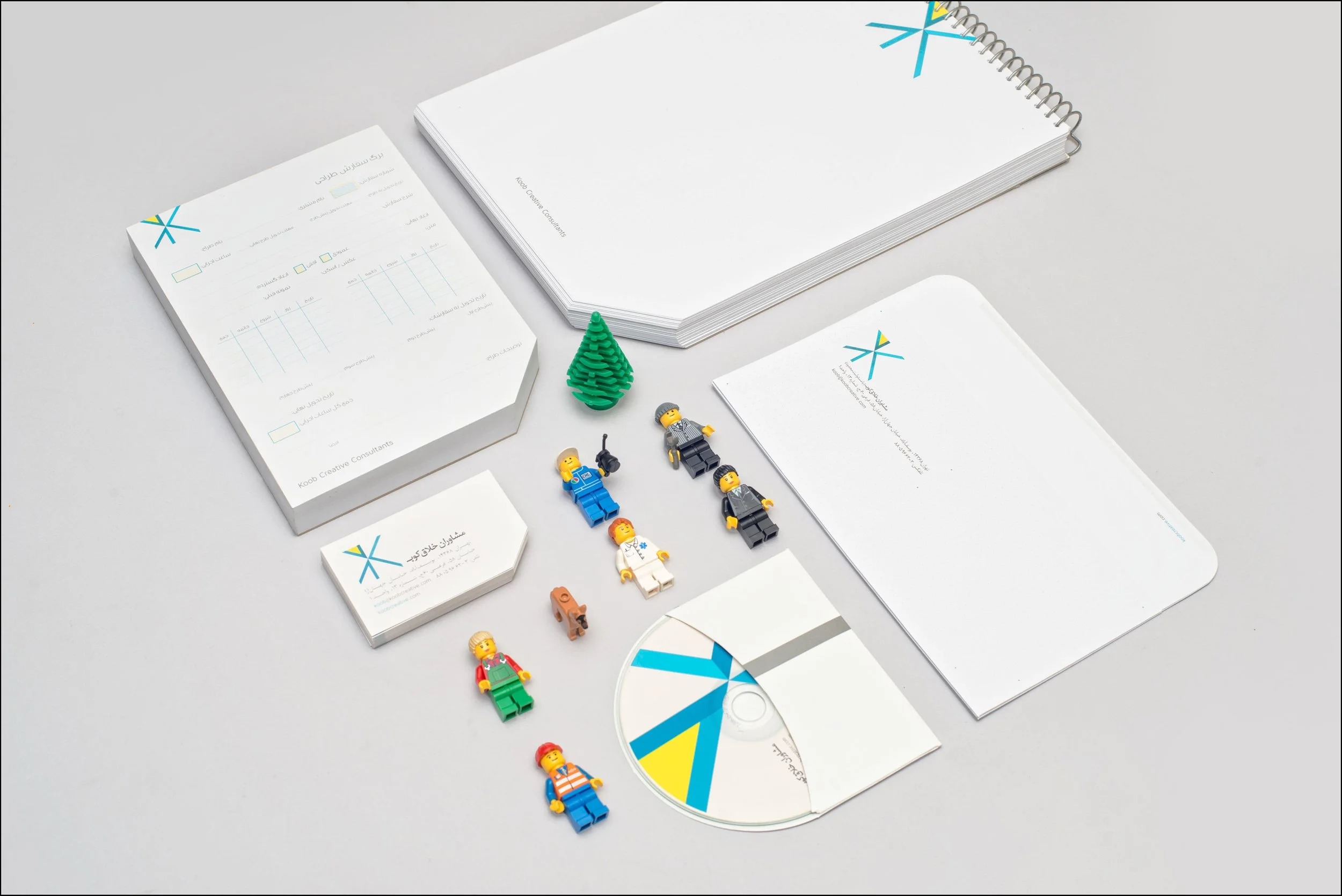 Open notebooks, business cards, miniature LEGO figures, a small LEGO tree, and a CD with a geometric design on the disc are arranged on a light gray surface.