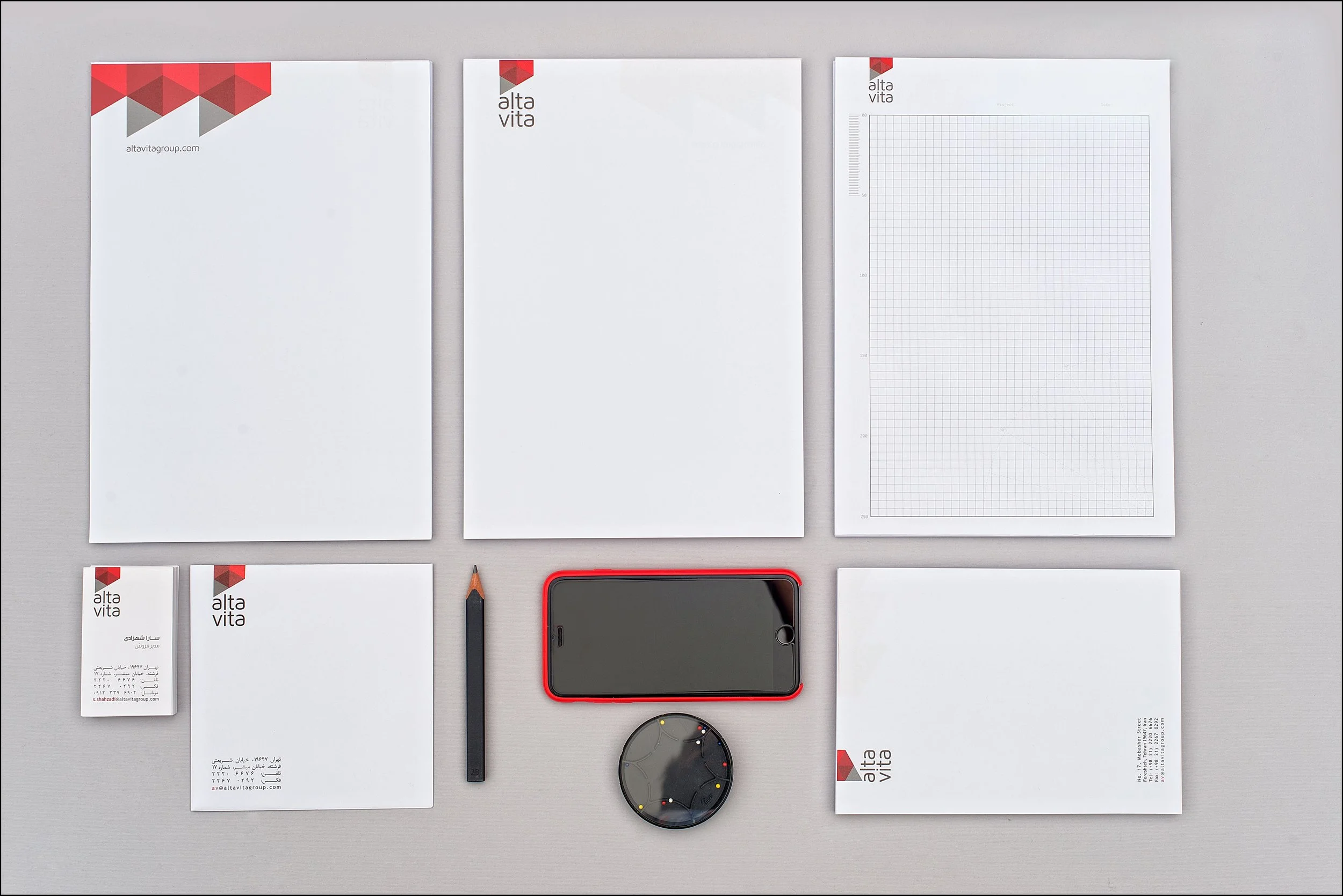 Office stationery including letterhead paper, notepads, a business card, a pencil, a smartphone with a red case, and a round paperweight, all branded with the 'alta vita' logo on a grey background.