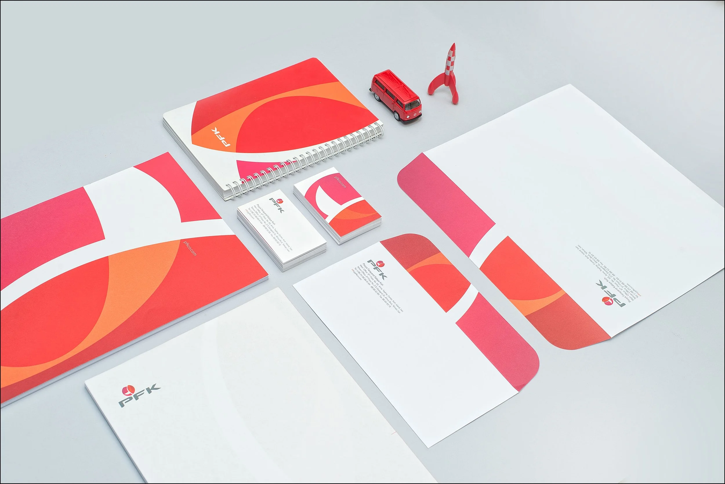 Branding stationery with items in red, orange, and white, including notebooks, business cards, letterhead, and folder, with a small red model bus and rocket on a gray surface.