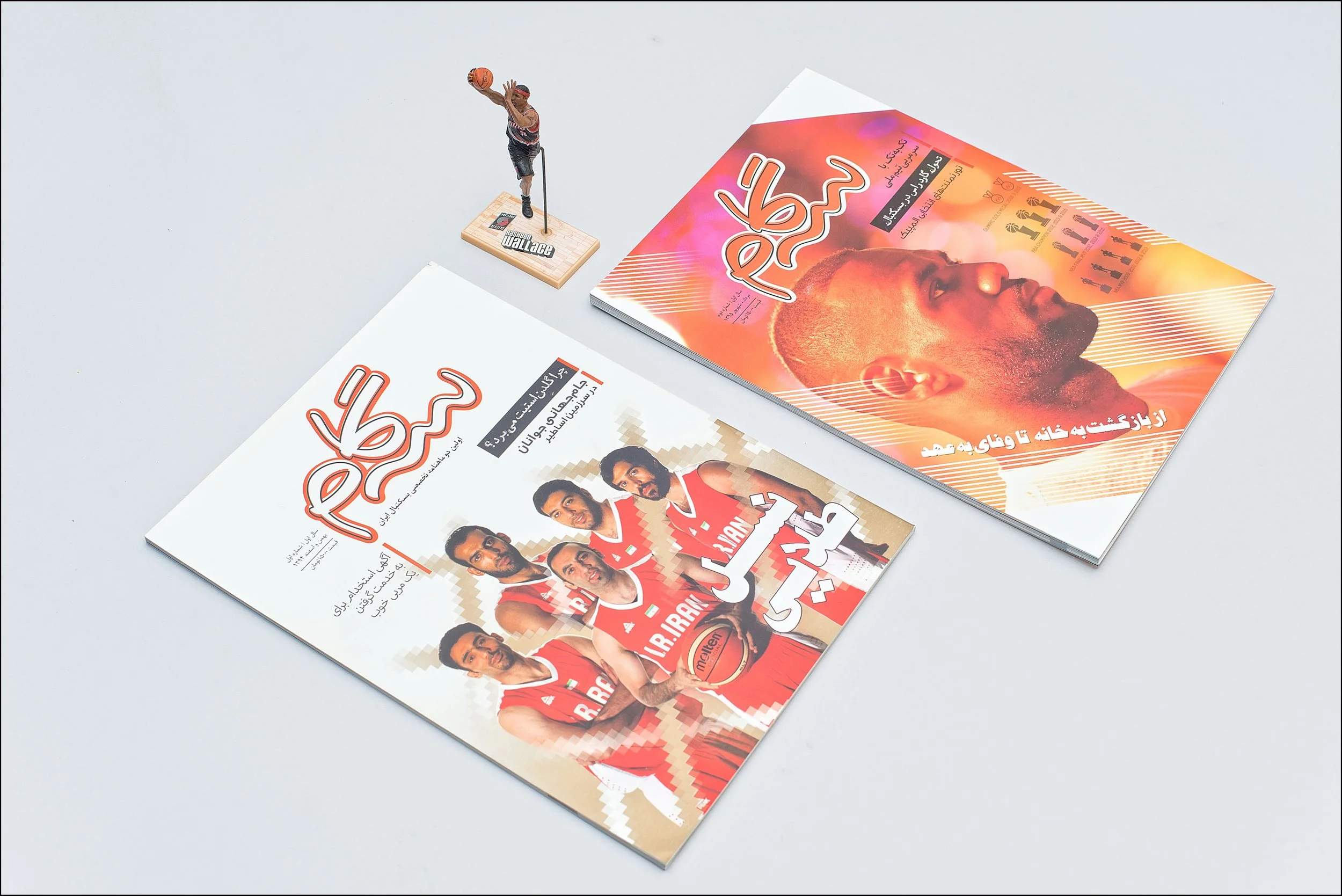 Two magazines with sports coverage and a basketball player figurine on a white surface. One magazine features a row of basketball players in red jerseys, and the other has a large profile photo of a man with a vibrant background.