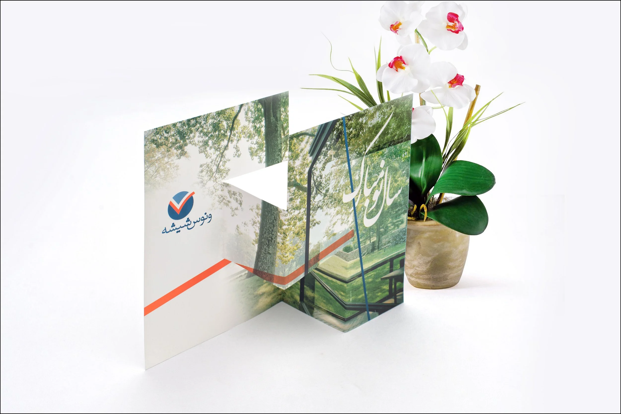 A three-panel brochure with an outdoor park scene and Arabic or Persian script, placed on a white surface with a potted orchid plant with white and pink flowers beside it.