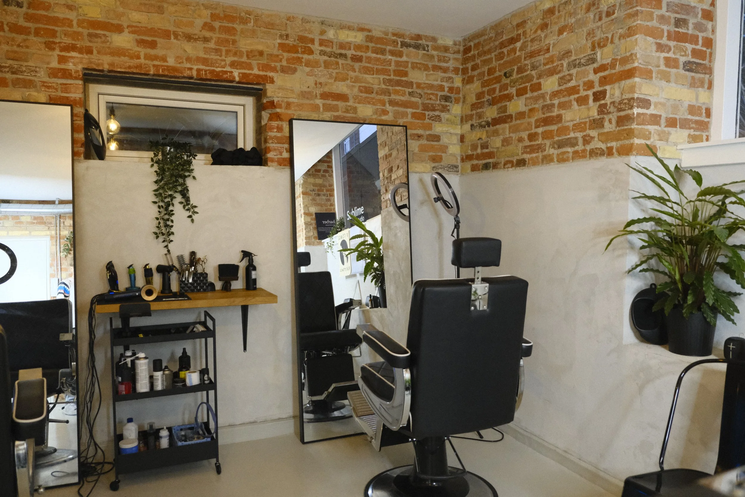 Barber shop interior with a black barber chair, brick walls, mirrors, a wooden shelf, hair styling tools, and potted plants.