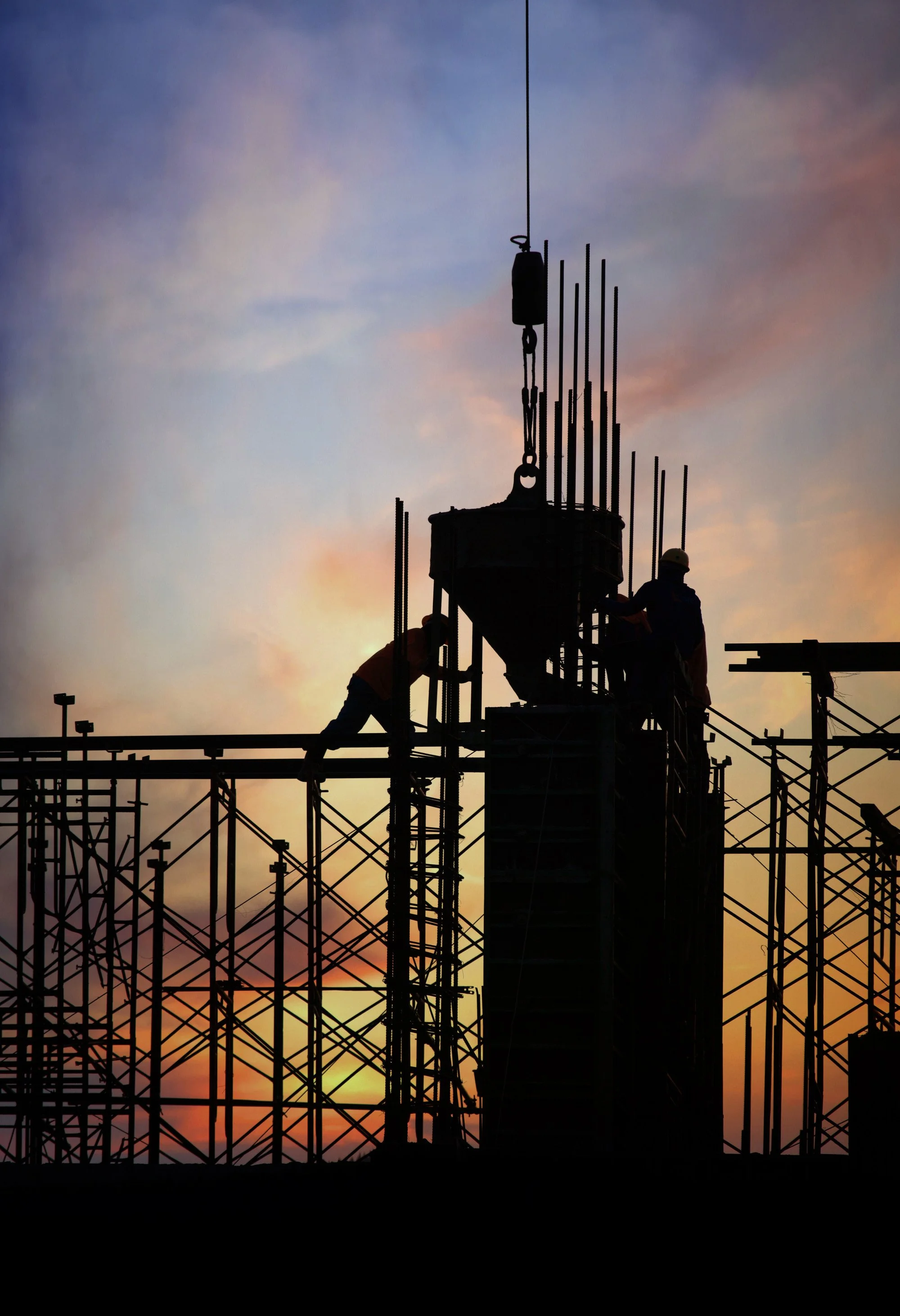 Silhouette di due operai edili che lavorano su una struttura di ponteggio al tramonto, con un cielo colorato di arancione, rosa e blu.