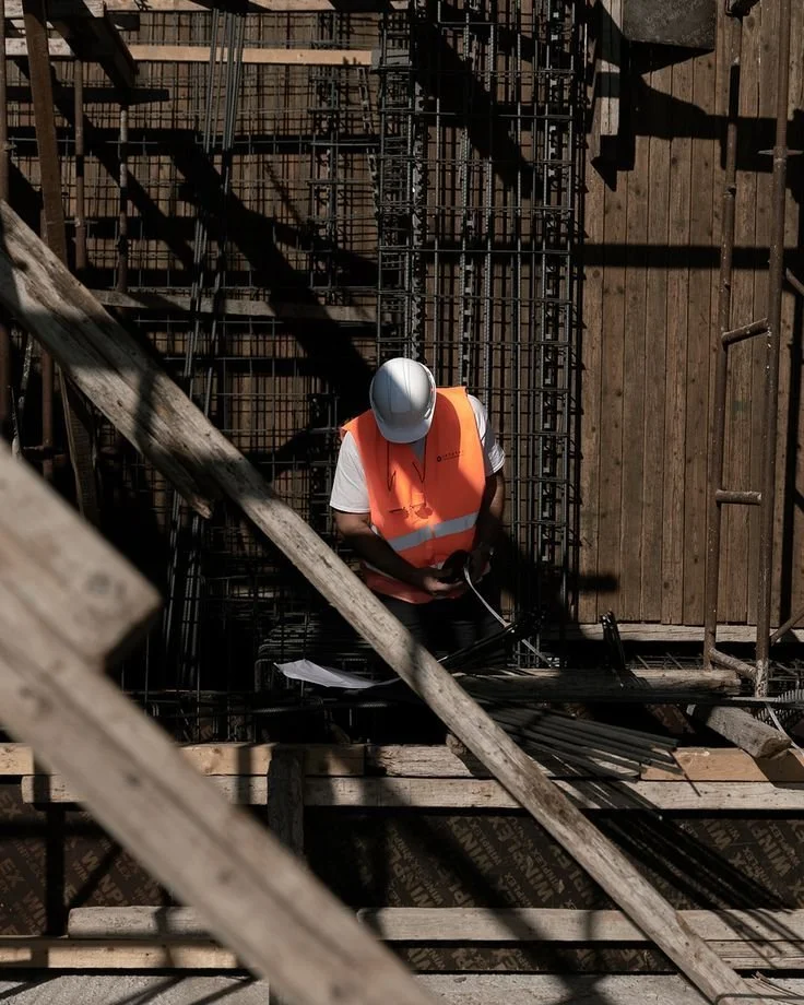 Impiegato al cantiere edile che indossa casco e giubbotto arancione, circondato da strutture in acciaio e legno.