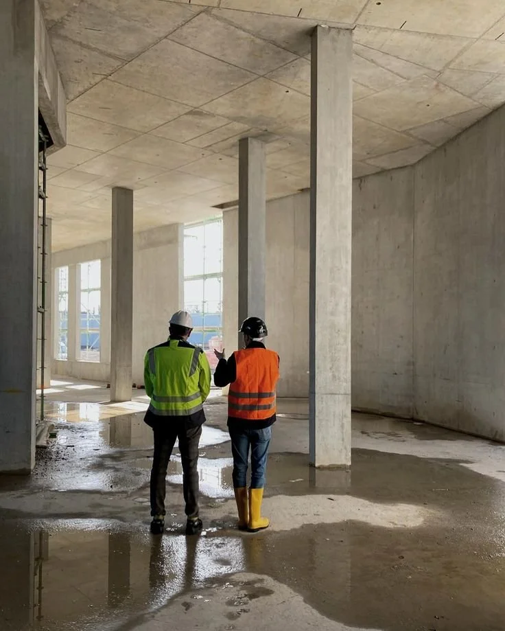 Due uomini con caschi e giubbotti di sicurezza discutono all'interno di un edificio in costruzione, con muri di cemento e grandi finestre, in un ambiente umido.