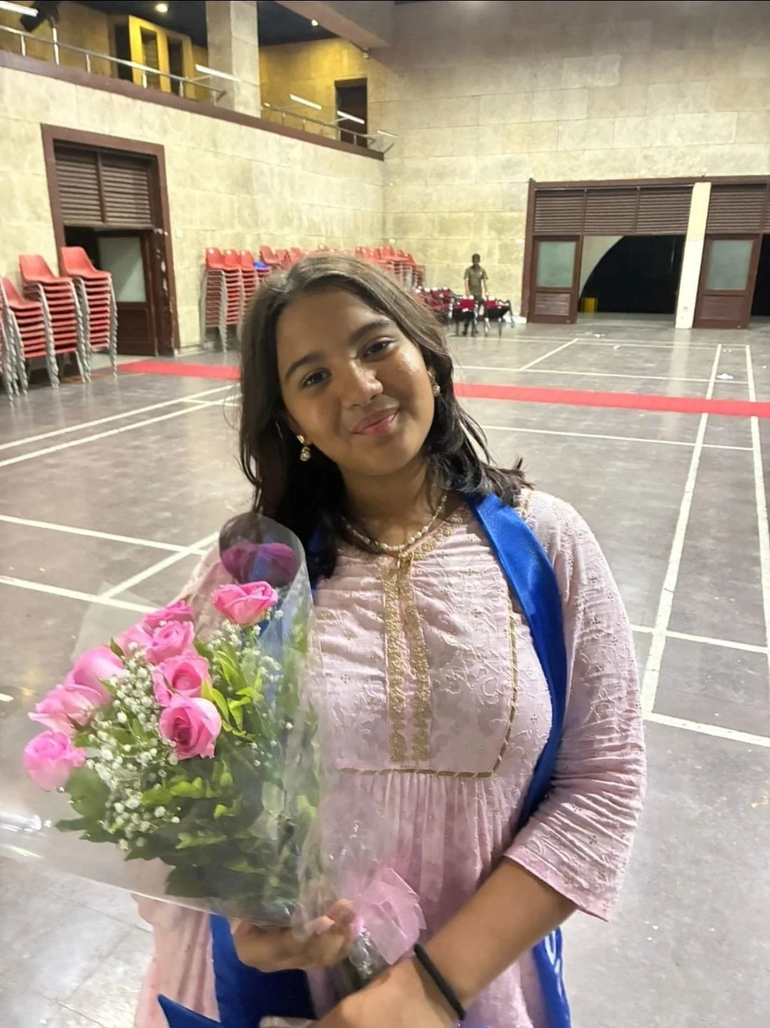 A young woman with black hair, wearing earrings, a necklace, and a light pink dress with gold embroidery, is holding a bouquet of pink roses wrapped in clear plastic. She is standing indoors in a large hall with chairs stacked against the wall and a man in the background.