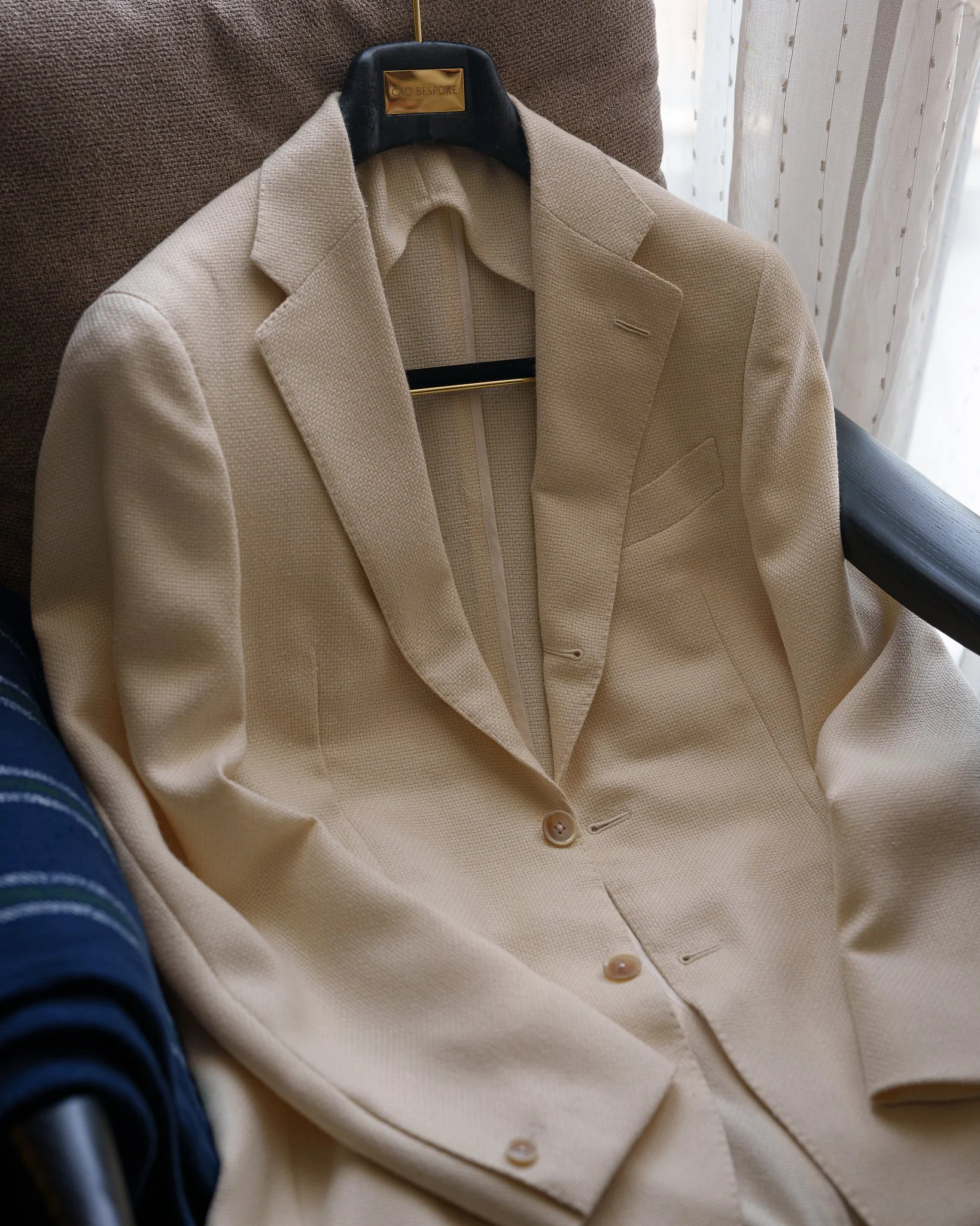 A beige suit jacket hanging on a black hanger on a brown armchair, next to a window with sheer curtains.