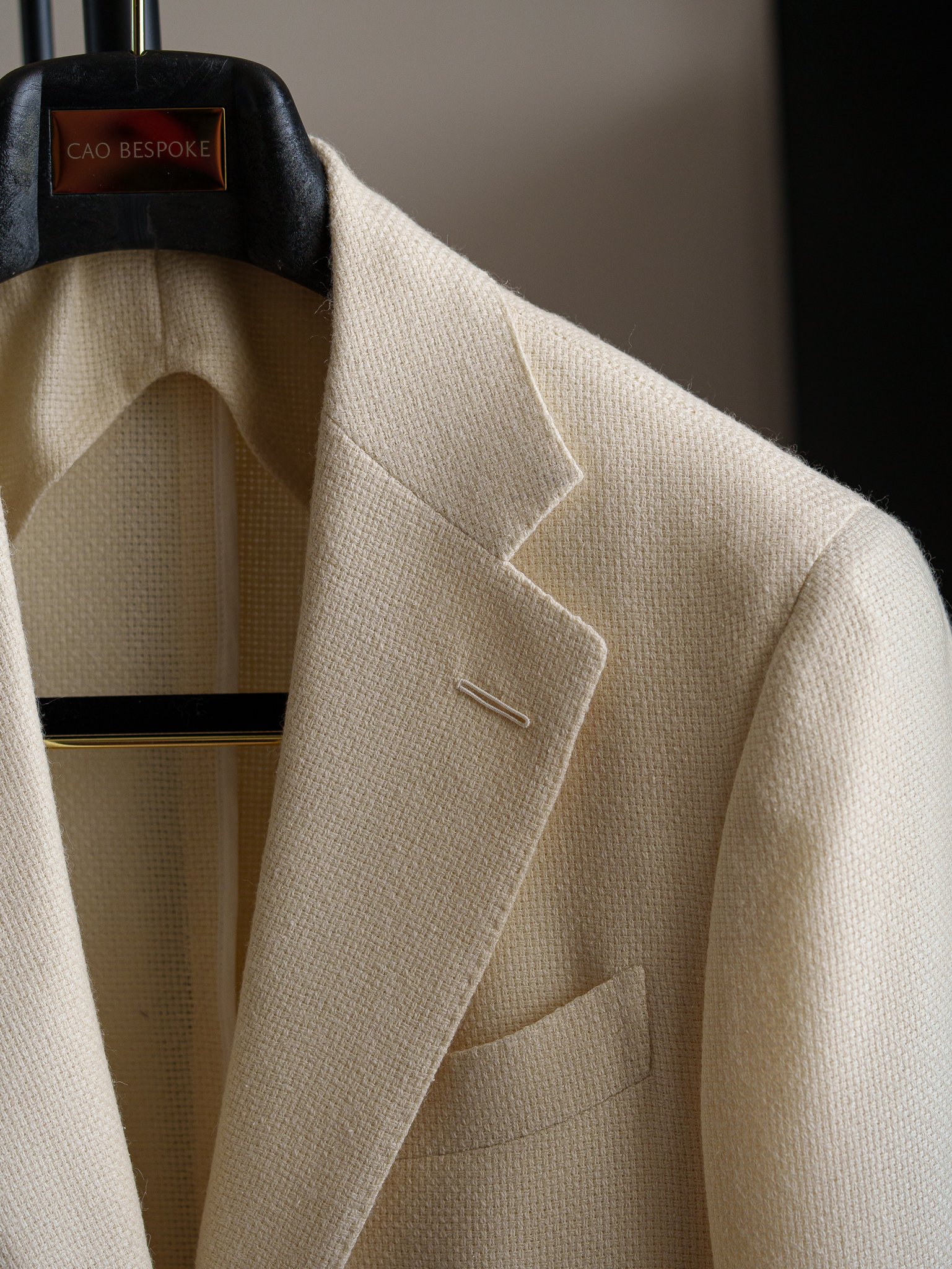 Close-up of a beige textured blazer with a notch lapel, hanging on a black hanger labeled 'Cao Bespoke'.