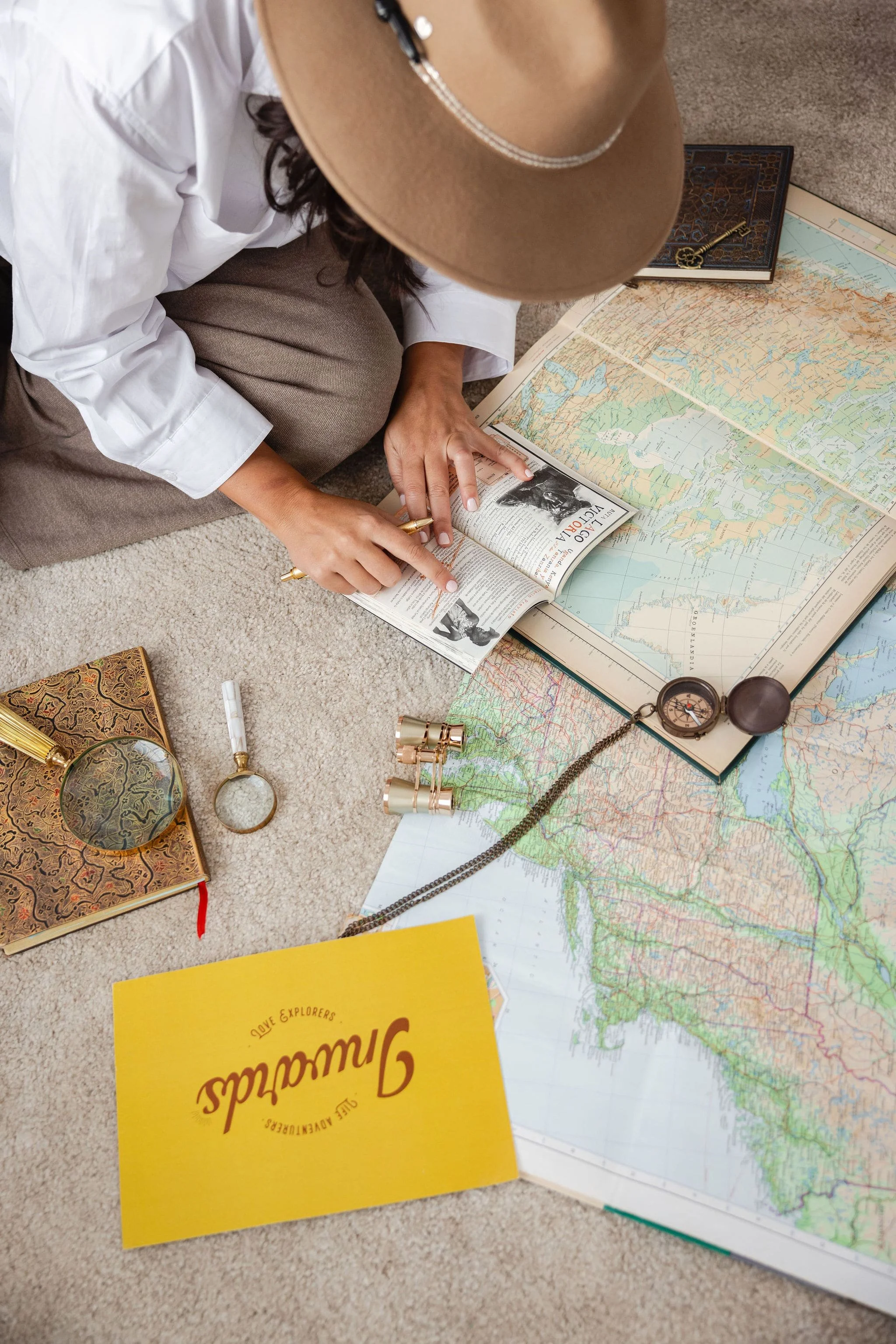 Mujer sentada frente a mapas, journal, brújula, binoculares y una tarjeta de INWARDS, representando la planificación simbólica de un viaje interior guiado por el autoconocimiento.