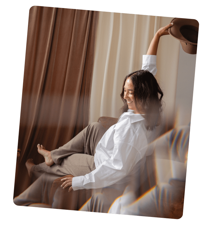 Mujer sonriendo en un sofá, sosteniendo un sombrero, vestido con blusa blanca y pantalones beige, en un ambiente acogedor con cortinas marrón y beige.