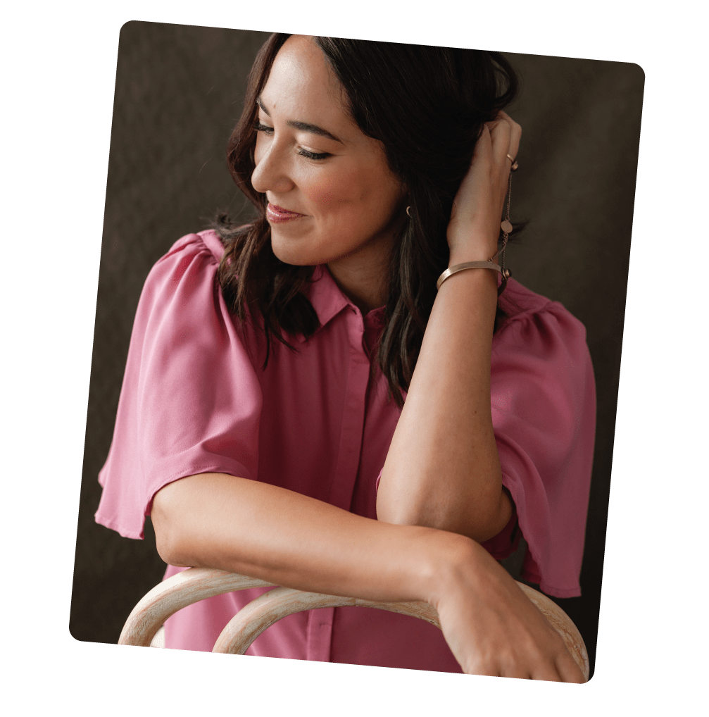 Mujer sonriente con blusa rosa, con cabello color café oscuro, tocándose la oreja, sentado en una silla de madera, fondo oscuro