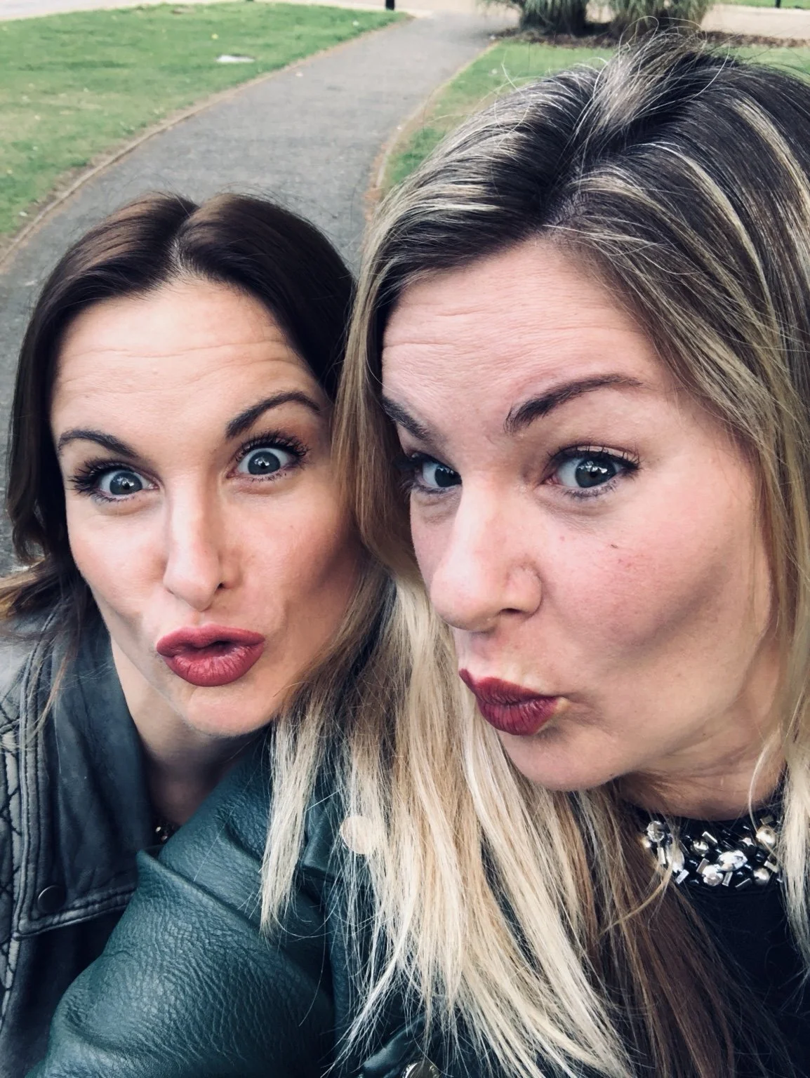 Two women taking a selfie outdoors on a paved walkway with green grass and bushes in the background.
