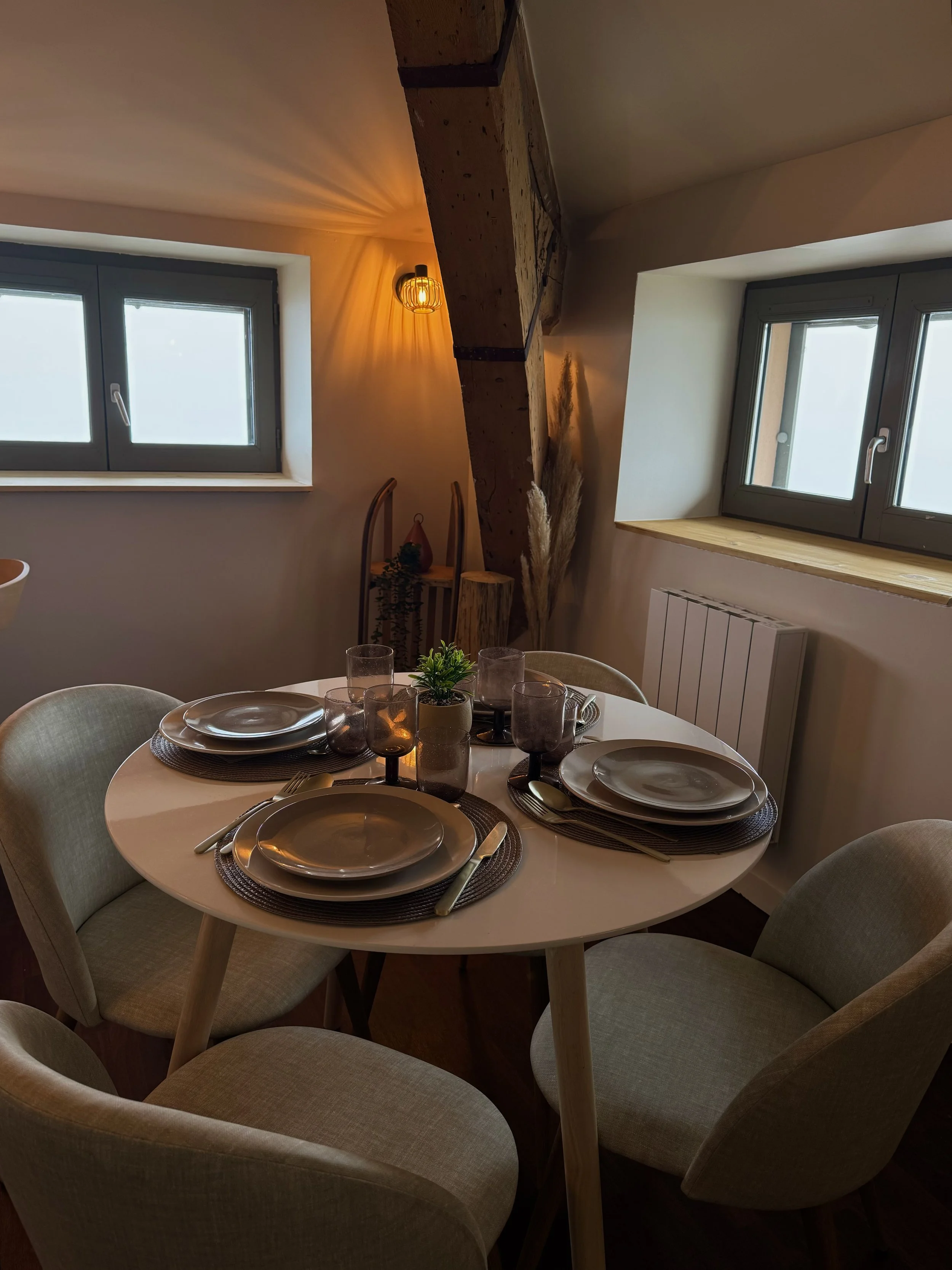 Salle à manger avec une table ronde dressée pour quatre personnes, assiettes, verres et couverts, dans une pièce chaleureuse avec fenêtres, un éclairage d'ambiance, et un décor en bois.