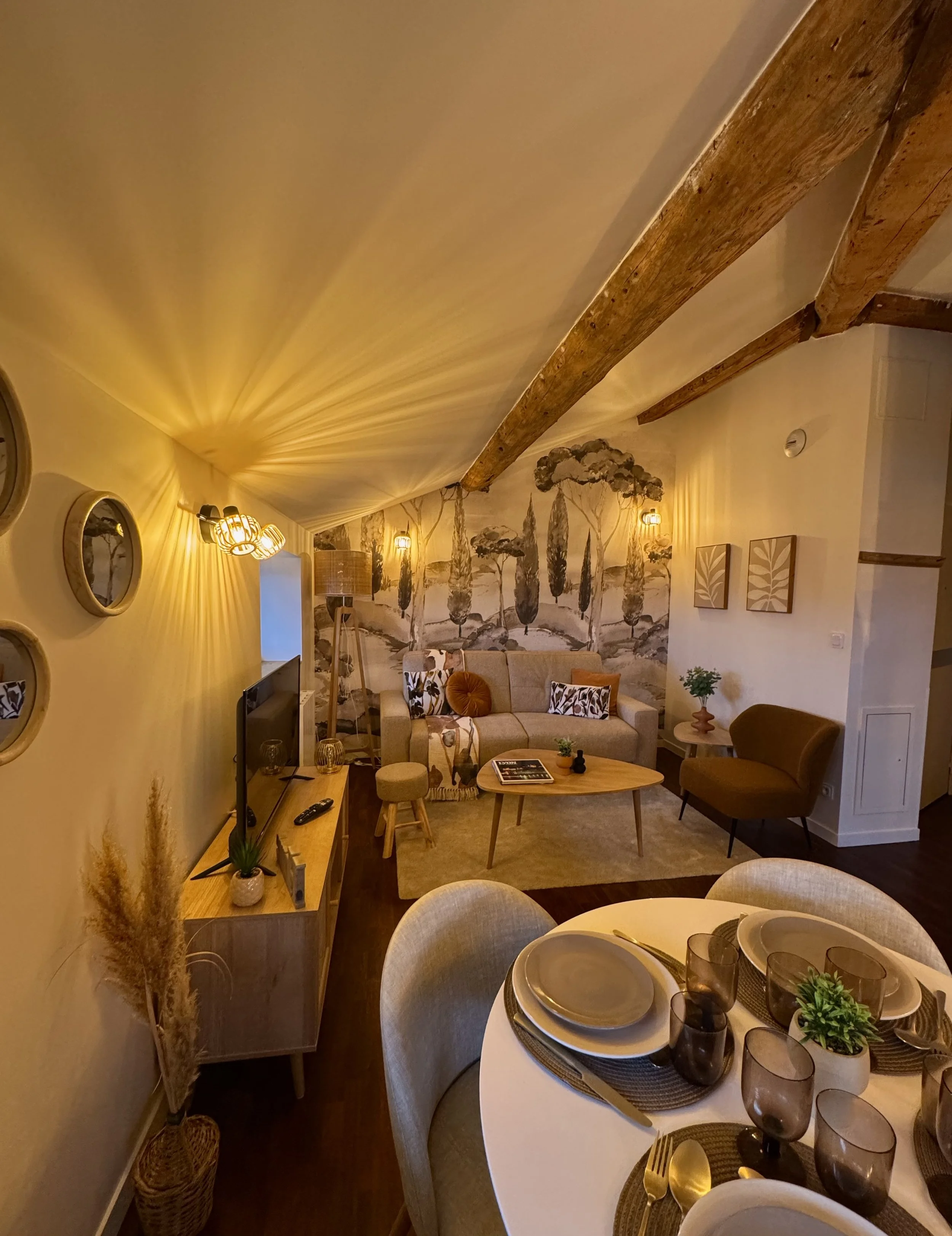 Salon et salle à manger dans un intérieur chaleureux avec mobilier en bois, canapé beige, fauteuil marron, tapis, mural avec peinture de paysages, table ronde avec vaisselle, verre et plantes décoratives, éclairage tamisé.