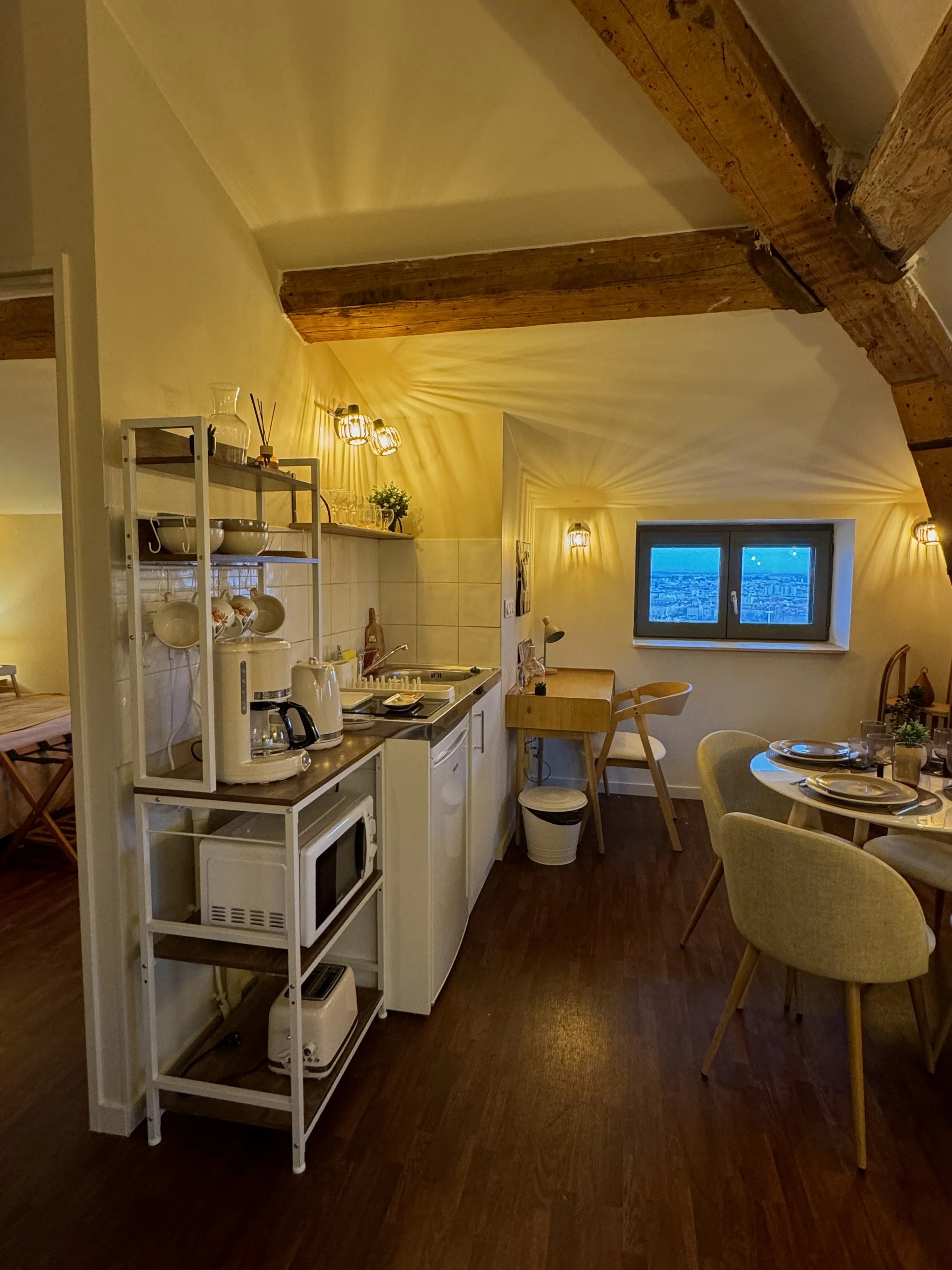Cuisine chaleureuse avec poutres en bois, table à manger dressée, lumière tamisée, fenêtre avec vue nocturne sur la ville, étagère blanche avec appareils électroménagers.
